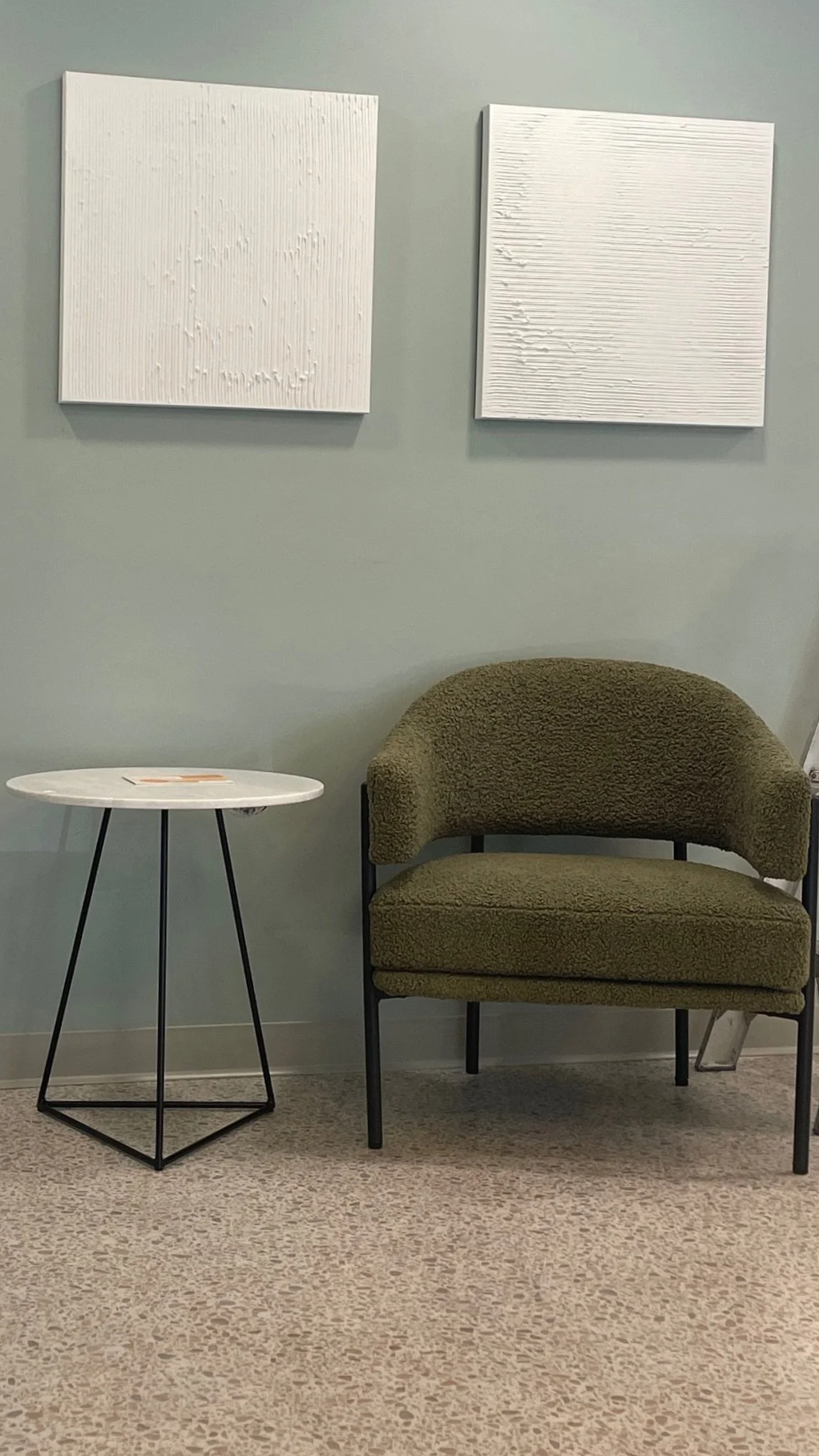 A waiting area with a green upholstered armchair, a small round white table with black metal legs, and two white textured wall art pieces on a light green wall.
