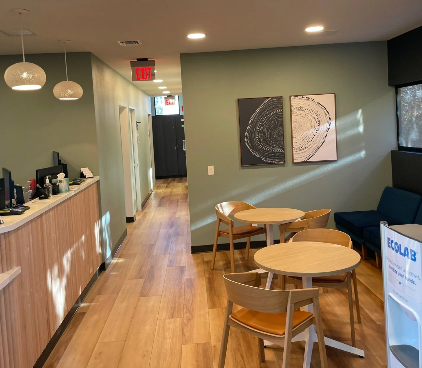 Interior of a modern office or waiting room with round wooden tables and chairs, a blue sofa, wall art, a front desk with multiple computer monitors, and a hallway with an exit sign.