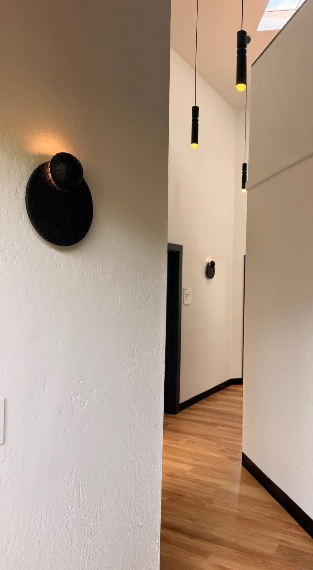 A modern hallway with white textured walls, mounted black wall art, black pendant lights hanging from the ceiling, wooden flooring, and a partially open door.