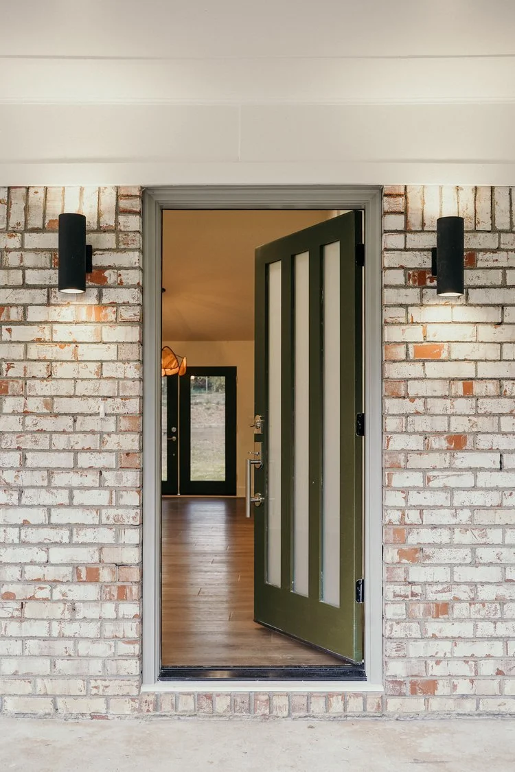 Exterior view of an open front door with a dark green color, showing a glimpse of the interior with wooden flooring and a black framed glass door inside.