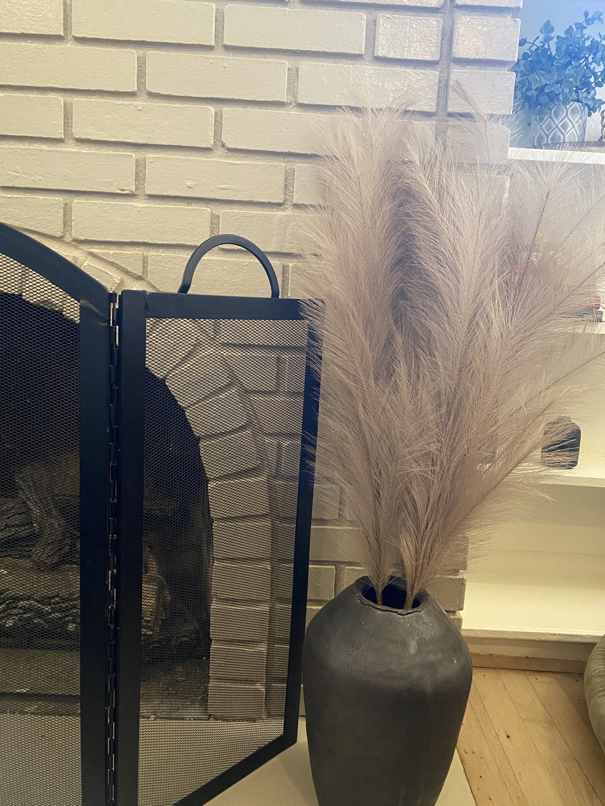 Large black vase with beige feathery ornamental grass in front of a white brick wall, partially obscured by a black metal fireplace screen.