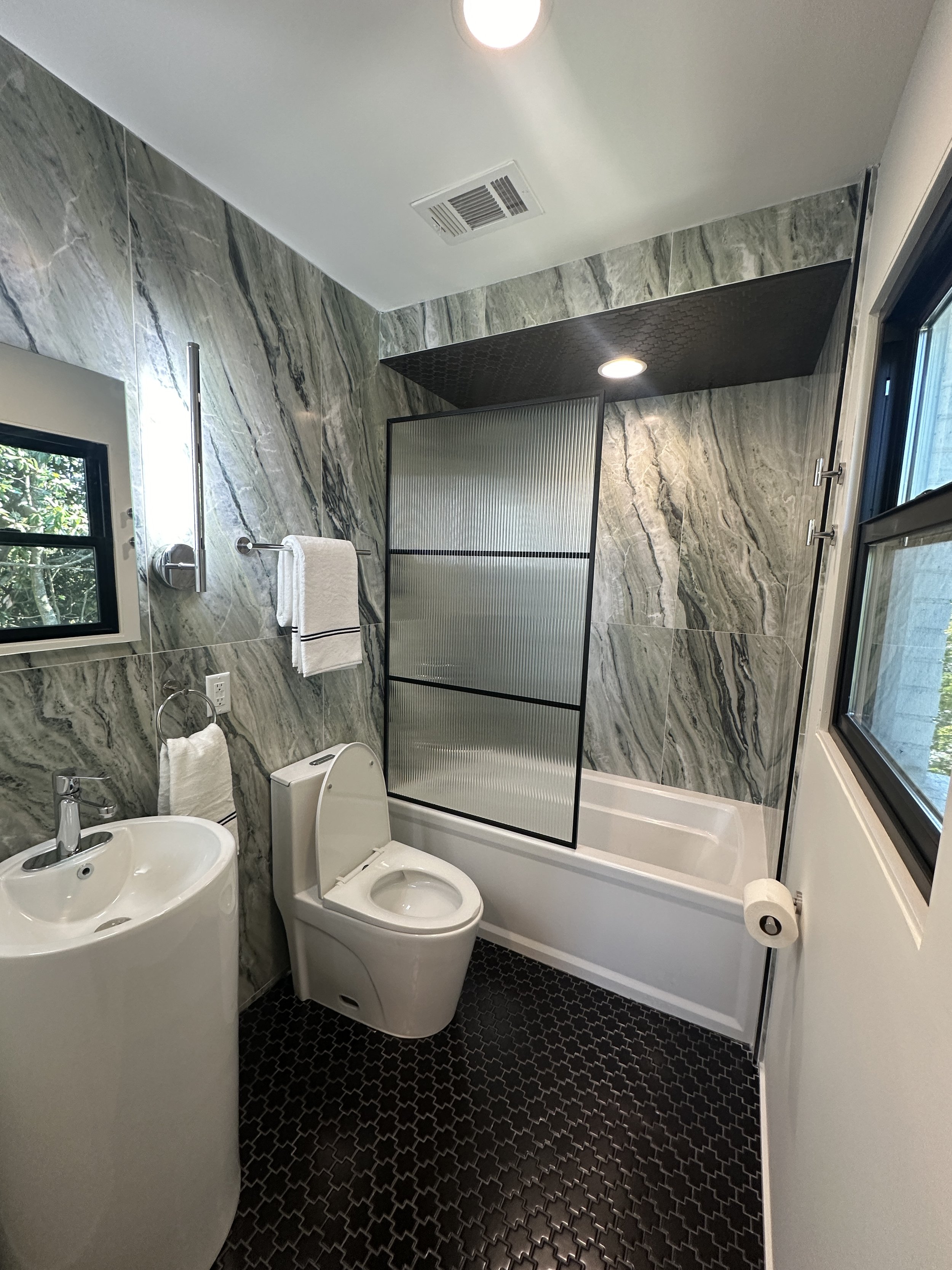 A modern bathroom with marble walls, black patterned floor tiles, a toilet, a bidet, a towel rack with a white towel, a window, a frosted glass shower partition, and ceiling lighting.