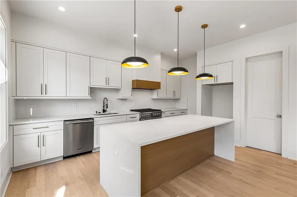 Modern kitchen with white cabinets, a large white island, pendant lights, stainless steel dishwasher, black faucet, and hardwood floors.