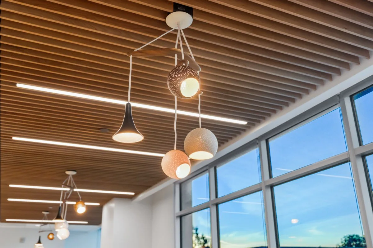 Modern hanging pendant lights with different shapes and colors in an indoor space with wooden slatted ceiling and large windows showing a blue sky.