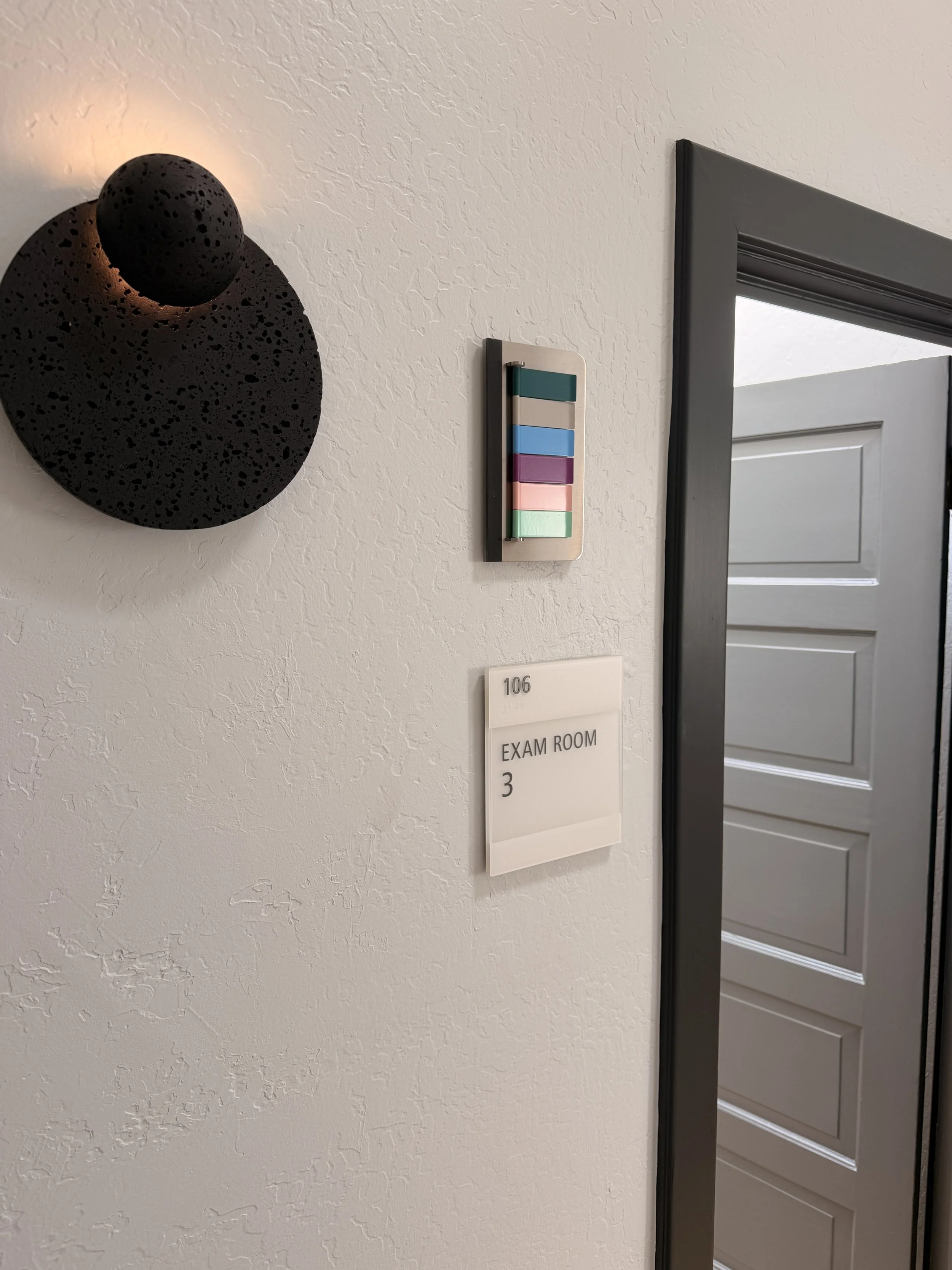 A wall with a textured white surface, decorated with a black spherical light fixture and a colorful wall-mounted board with horizontal pastel-colored sections. A sign indicates the room is an exam room number 3, next to a partially opened gray door.