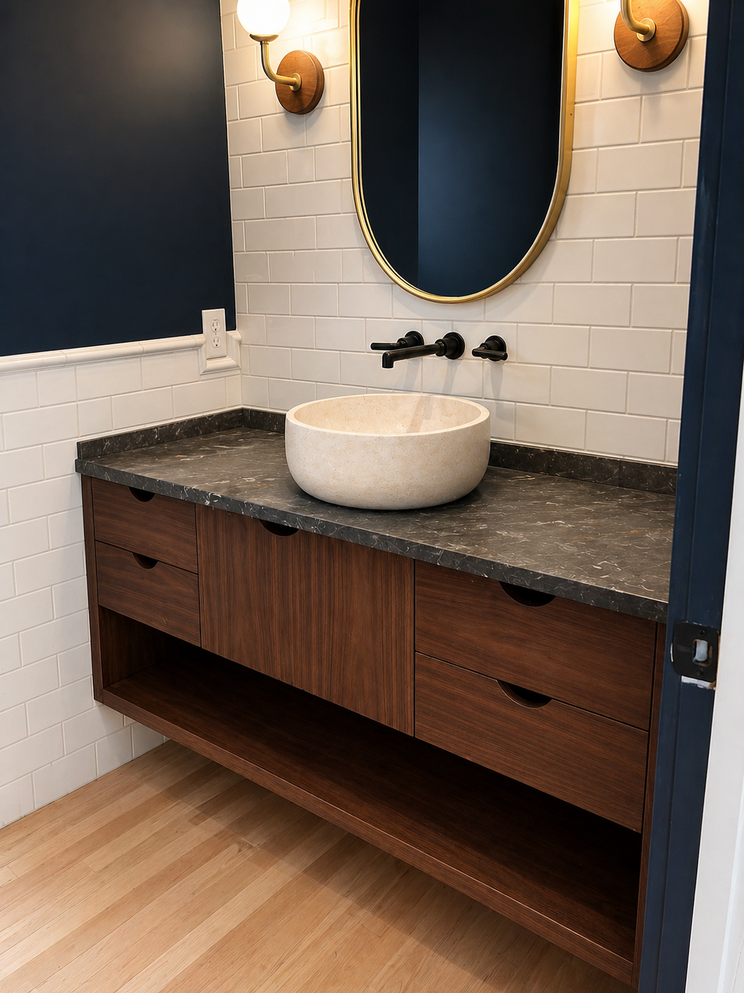 Bathroom vanity with a beige stone vessel sink, black faucet and wall-mounted light fixtures, mirror, and white subway tiles.