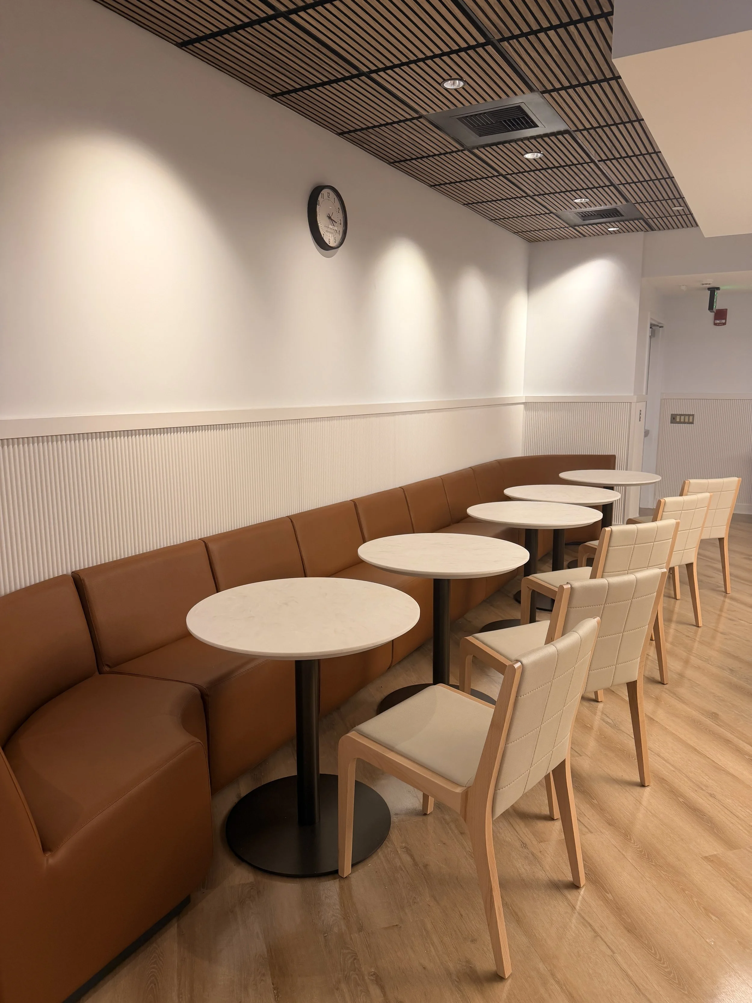 Empty cafe seating area with a brown curved sofa with white marble-top tables and beige chairs with wooden frames, cream-colored walls, a small wall clock, and a wooden ceiling with integrated ventilation vents and recessed lighting.