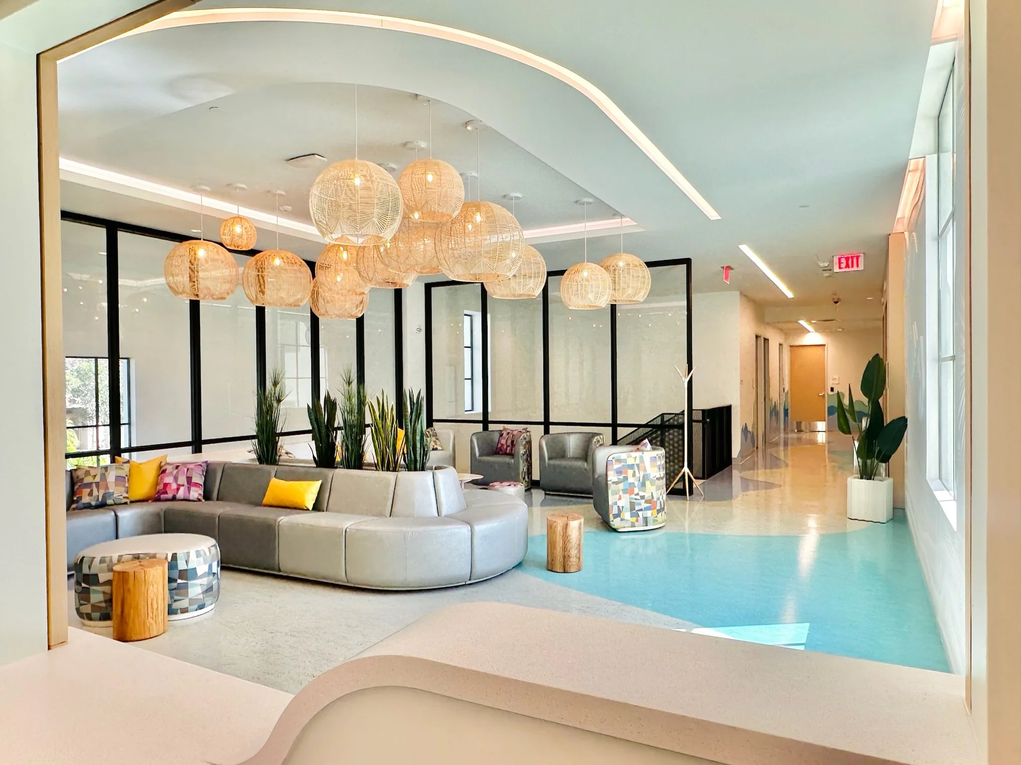 Modern hotel lobby with round gray sofa, colorful pillows, hanging wicker pendant lights, plants, and art decor. Large windows allow natural light, and a hallway leads to other areas.