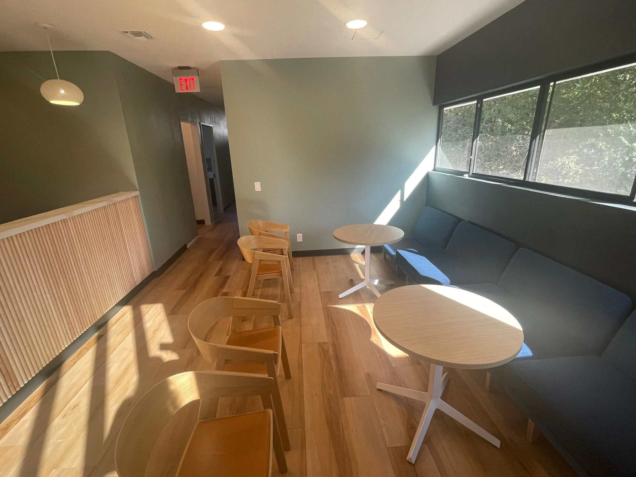 Interior view of a cafe or lounge with green walls, wooden flooring, a row of light-colored wooden chairs, small round tables, a gray cushioned seating area, a large window letting in natural light, and a red exit sign on the ceiling.