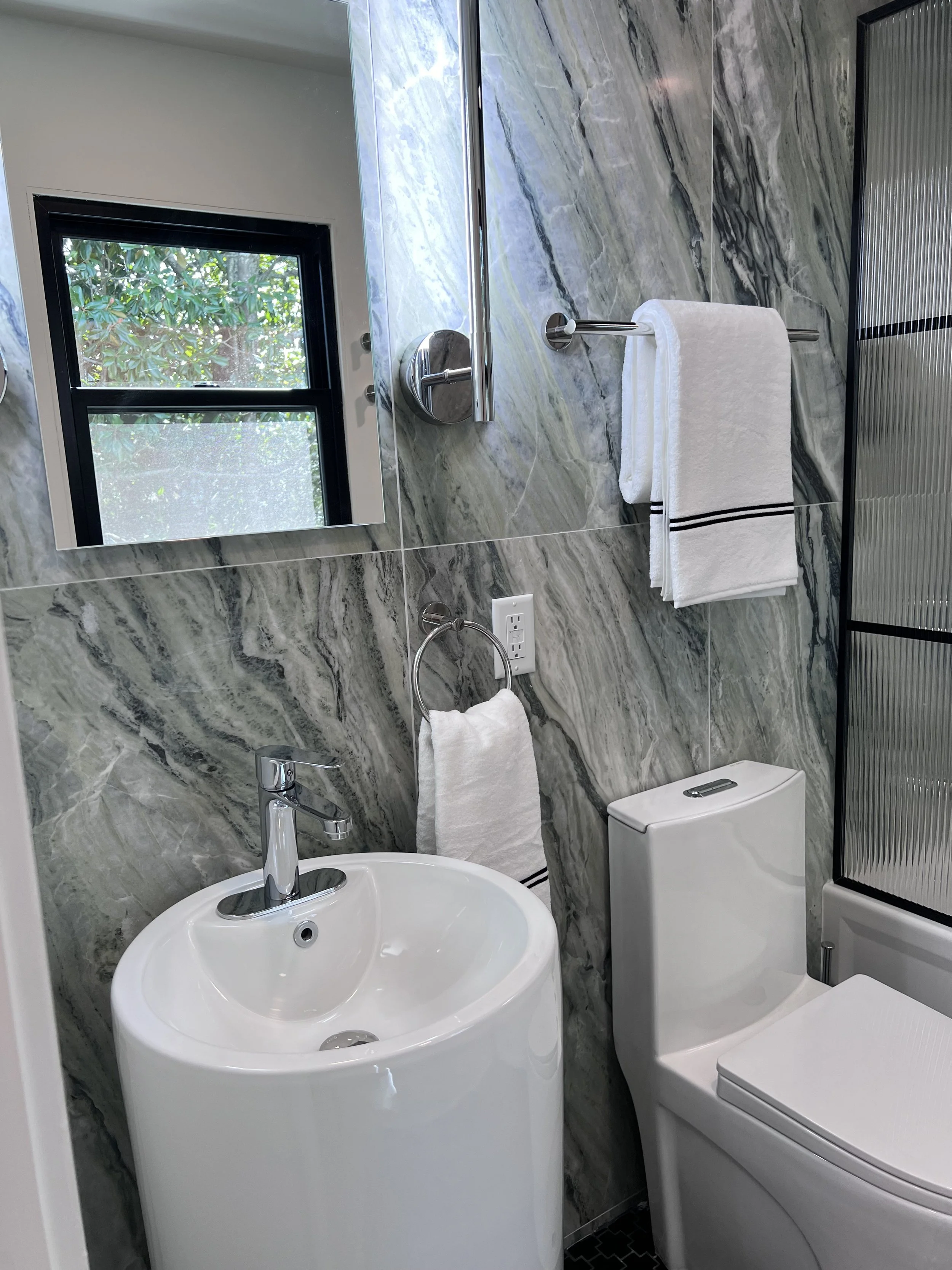 Modern bathroom with a small white sink, a round mirror, a toilet, a towel rack with a white towel, a window with greenery outside, marble walls, and a black and silver shower enclosure.