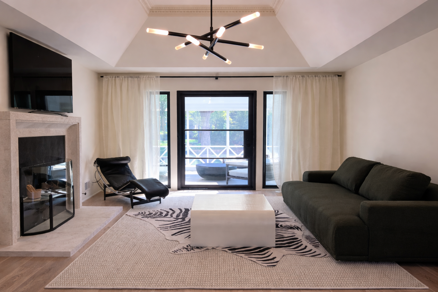 Living room with modern decor, black armchair, green sofa, white coffee table, fireplace, TV, doorway leading outside, white curtains, and a black ceiling light fixture.
