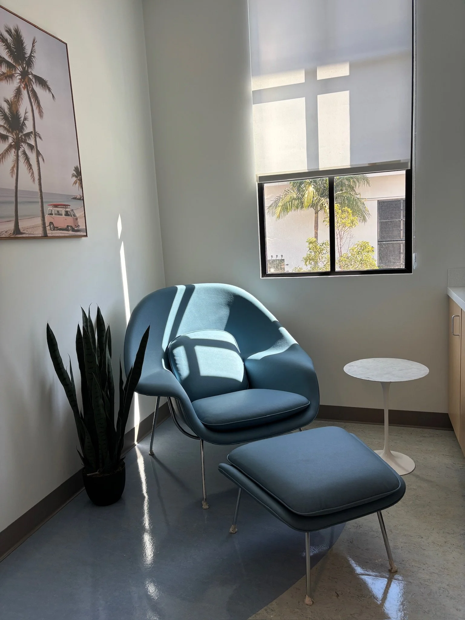 A modern waiting room with a blue lounge chair and footstool, a small white side table, a potted plant, a framed beach-themed poster on the wall, and a window with blinds showing palm trees outside.