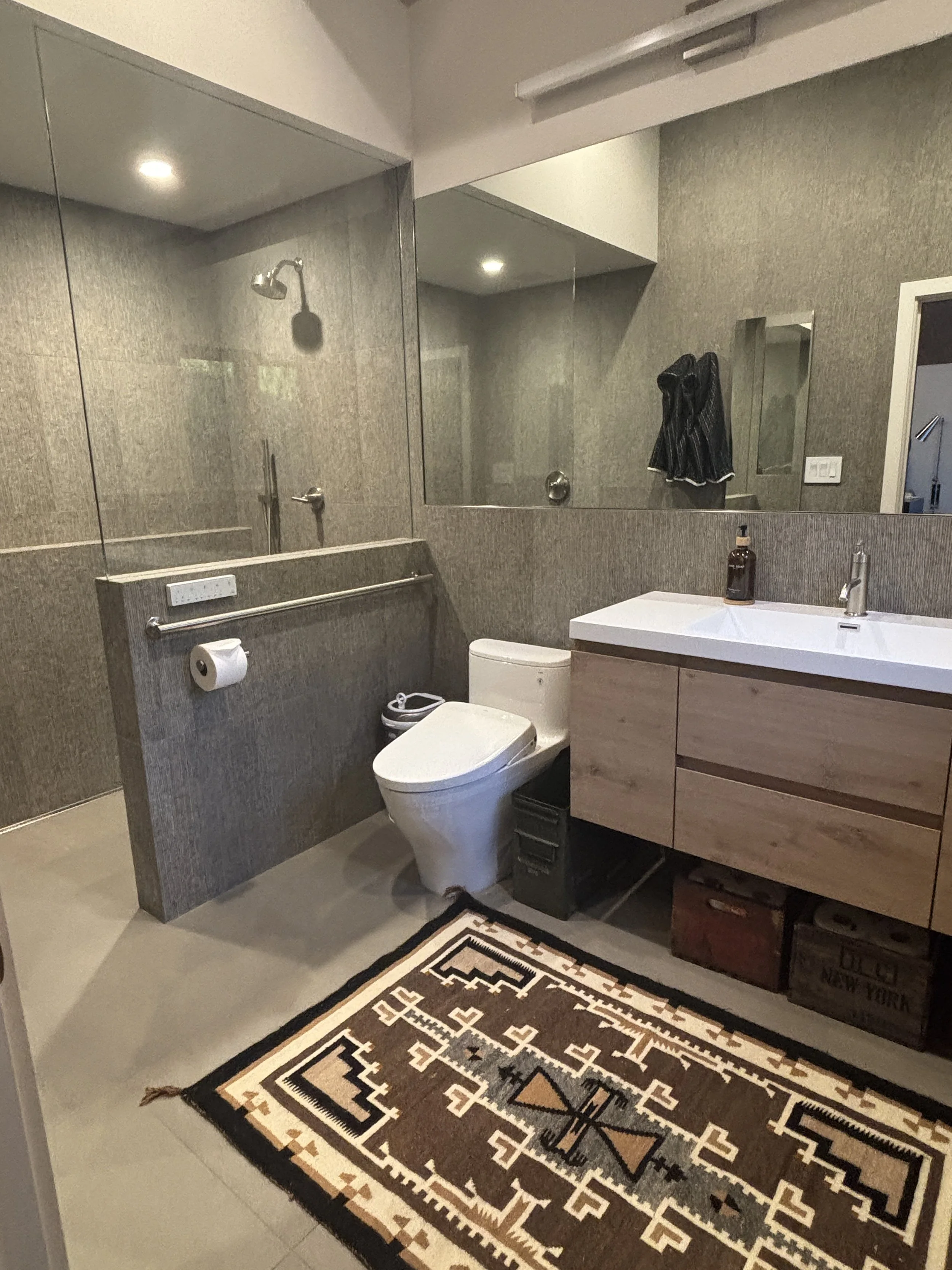 A modern bathroom with a walk-in shower enclosed by glass, a wall-mounted mirror, a white sink with a wooden cabinet, a toilet, a decorative rug on the floor, and a patterned towel hanging on the wall.