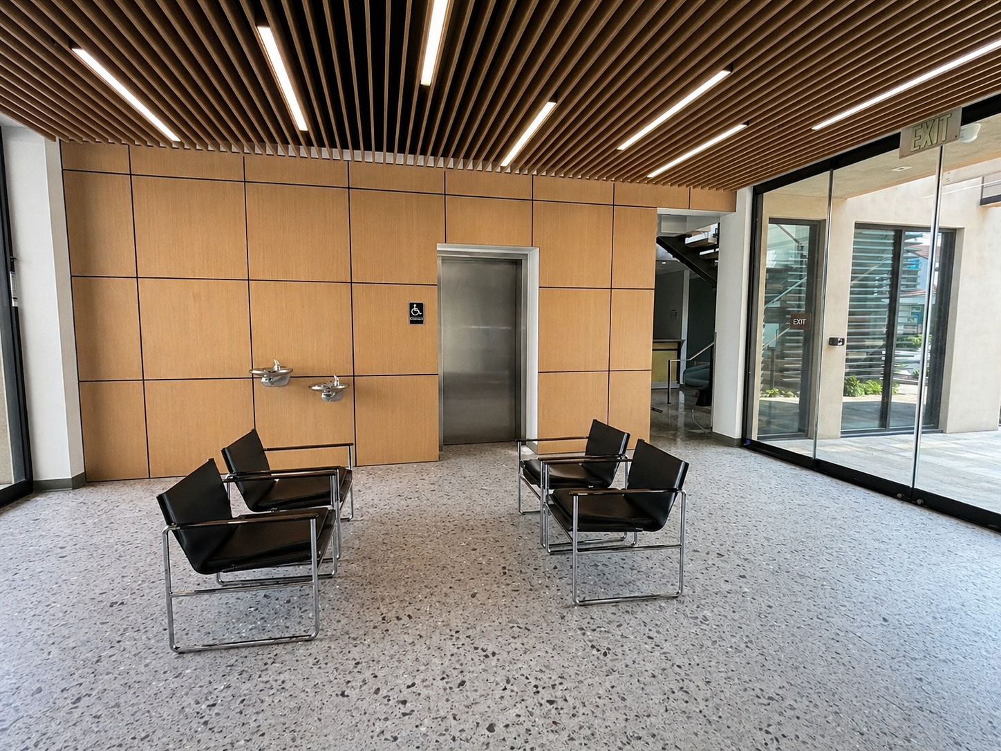 An indoor space with speckled flooring, four black chairs with metal frames, an elevator with a silver door, a wheelchair-accessible sign, two water fountains, large glass doors, and wooden wall paneling.