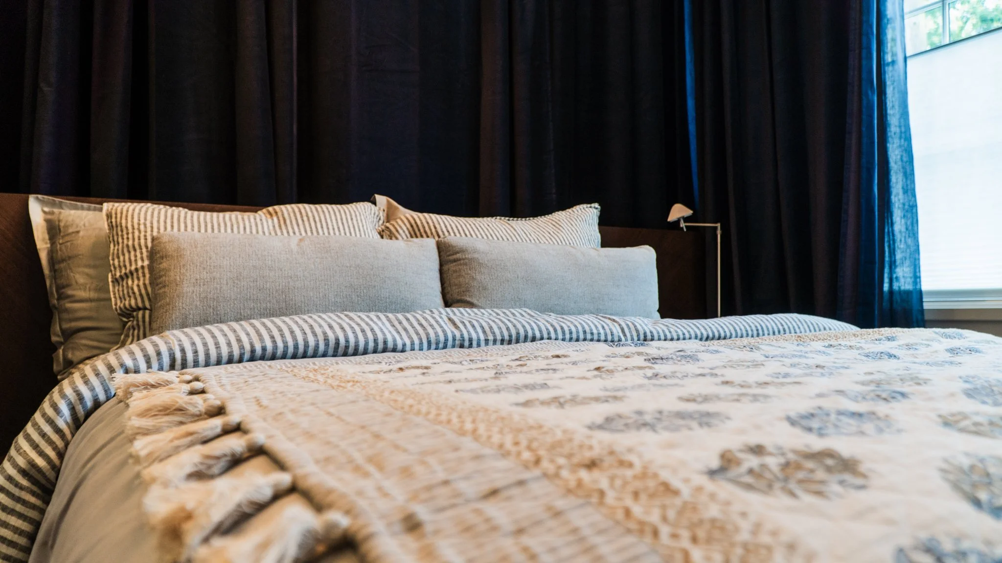 A neatly made bed with multiple pillows and a bedspread, set against a dark curtain and illuminated by natural light from a window.