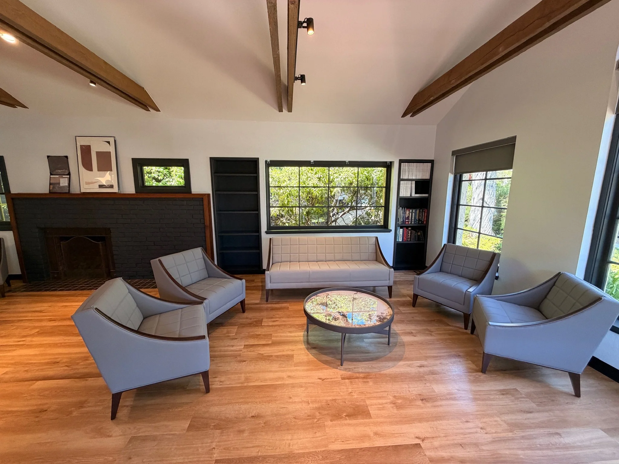 Living room with wooden floor, multiple light gray chairs, a glass topped coffee table, large windows with black frames, a built-in black bookshelf, and a fireplace with a dark brick surround.
