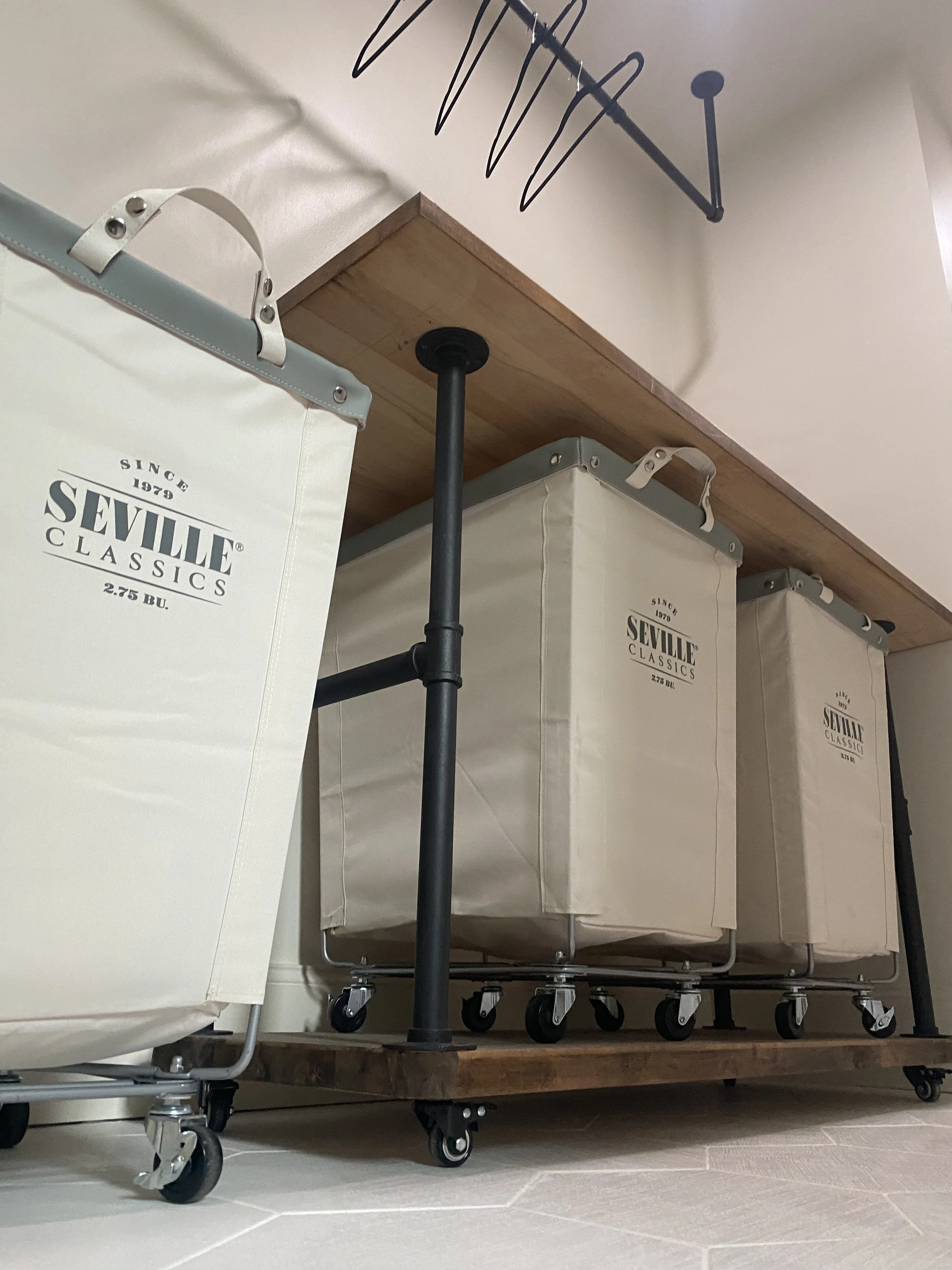 A home laundry room or closet with laundry baskets on a wheeled cart, white fabric with label 'Seville Classics' hanging from a wall hook, and hanging storage above.