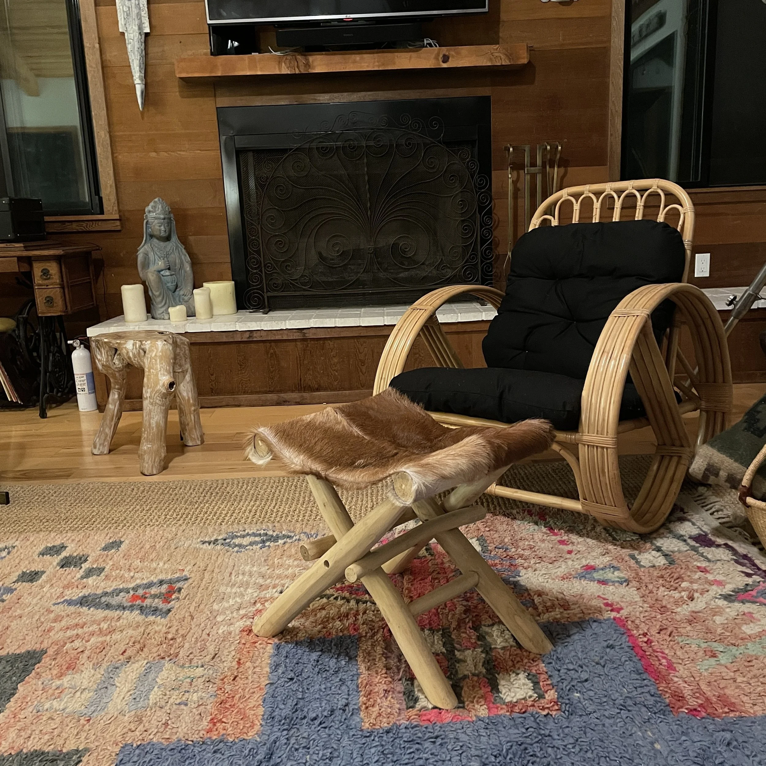 A room with wooden walls and a multicolored woven rug. A wicker chair with a black cushion, a small wooden table, and some candles. A gray statue of a woman is on the table. There is a fireplace with a decorative metal screen, a television above, and