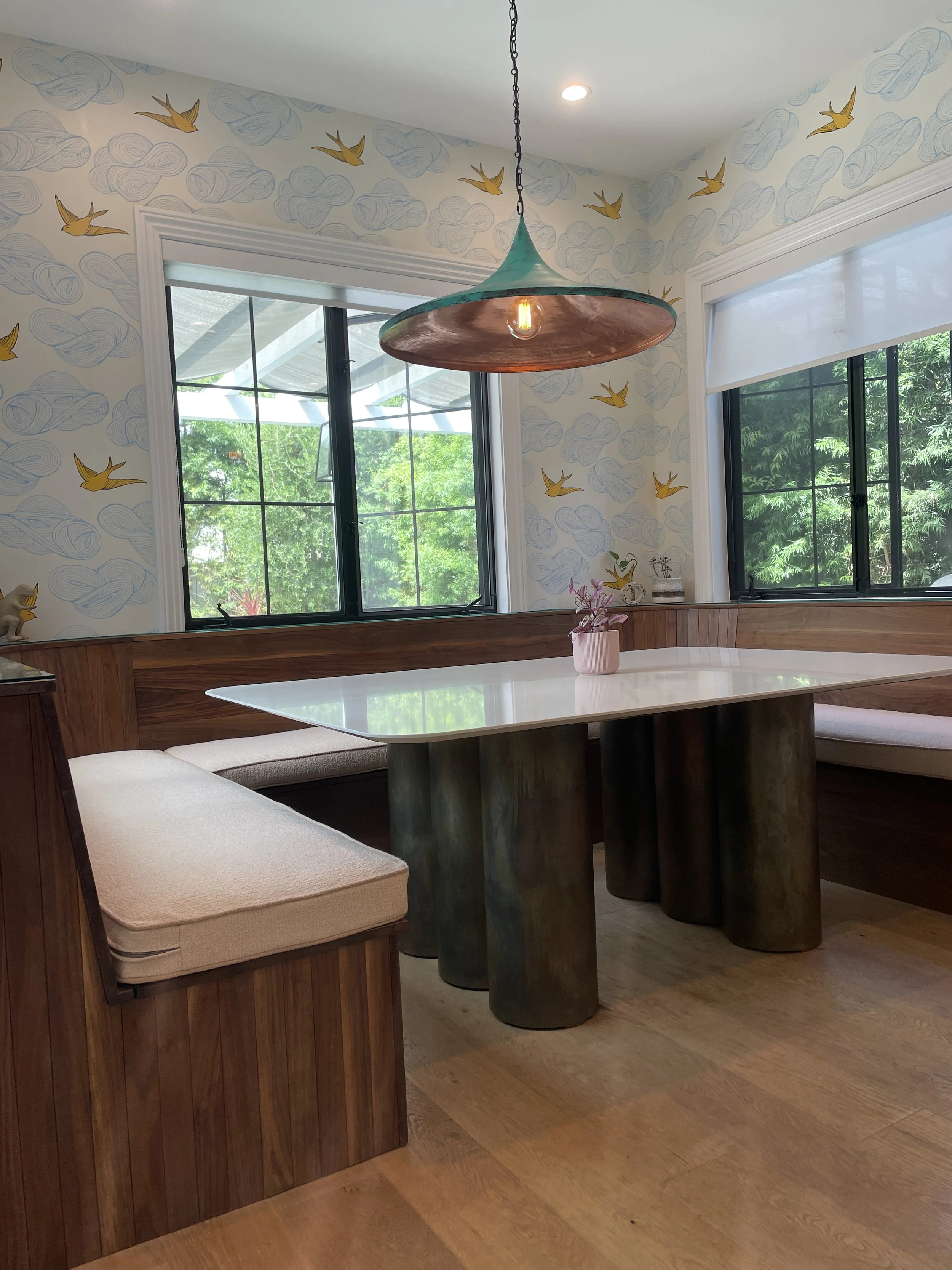 Dining nook with a white table, built-in wooden bench with beige cushions, and a pink potted flower on the table. Two large windows with black frames show greenery outside, with patterned wallpaper featuring clouds and yellow birds. A copper-colored 