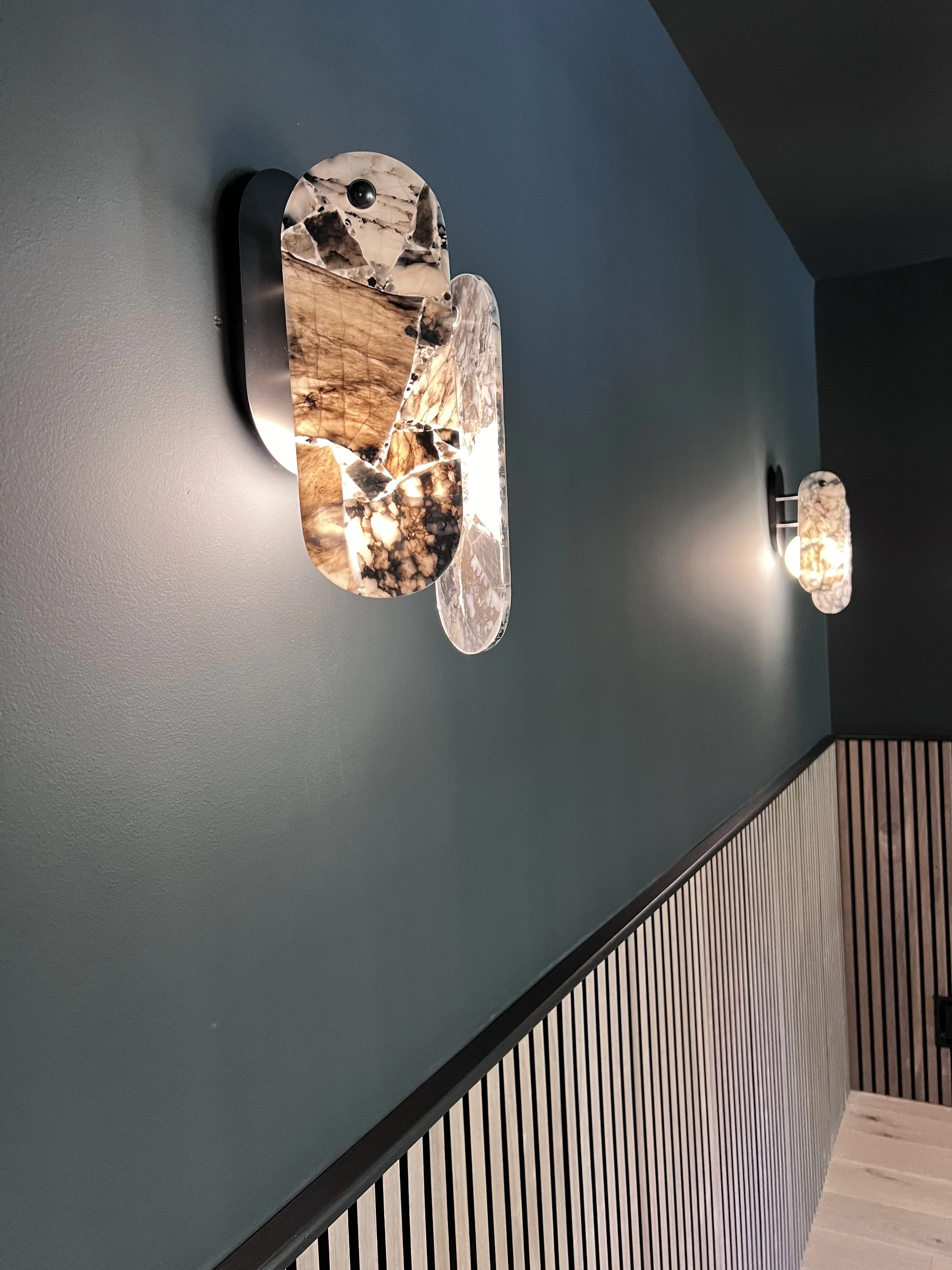 Modern interior lighting with marble and metal wall fixtures emitting warm light, dark teal wall, black and wooden slatted paneling.