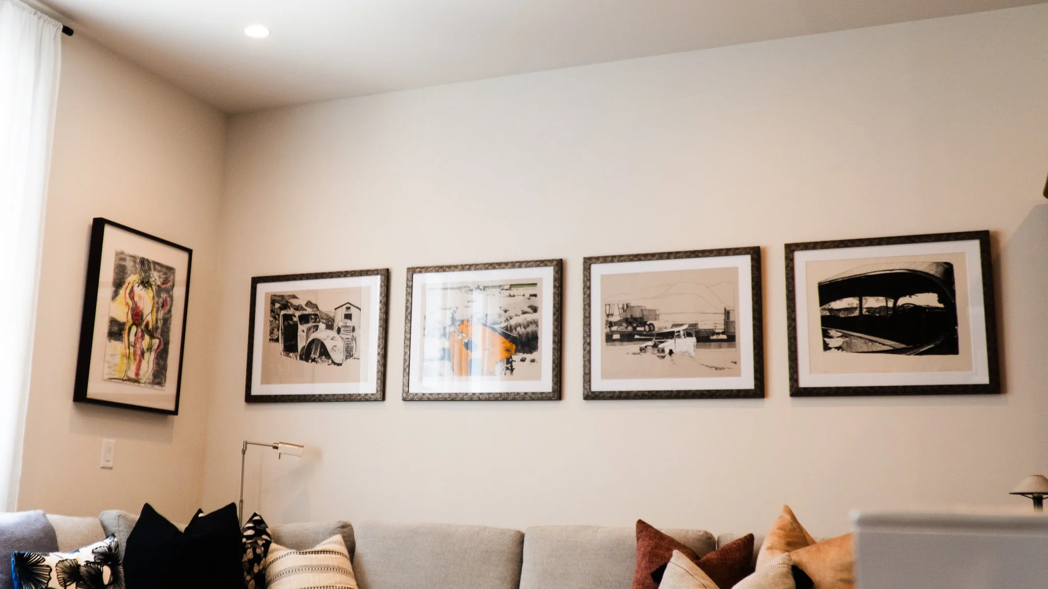 Interior of a living room with a white wall decorated with five framed black and white and colored artwork. Below, there is a beige couch with multiple pillows, including black, white, and brown ones.
