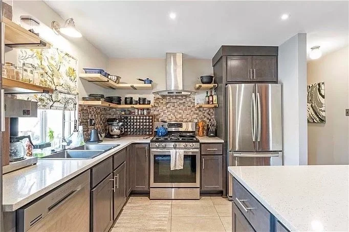 Modern kitchen with gray cabinets, stainless steel appliances, open shelving with dishes, and a patterned backsplash.