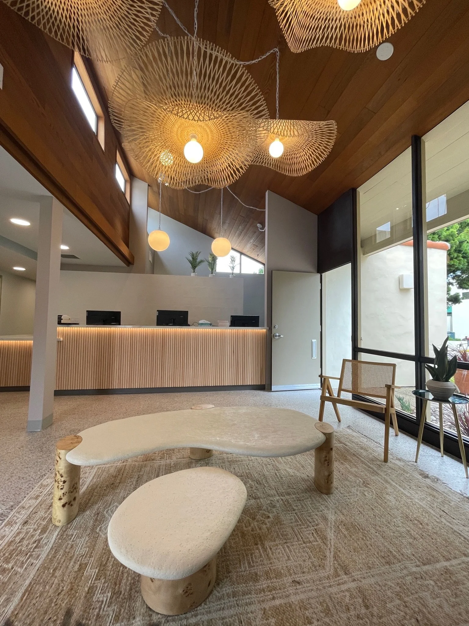 Modern interior space with wooden ceiling, decorative pendant lights, large windows, and minimalist furniture including a round stone table and a wooden chair.