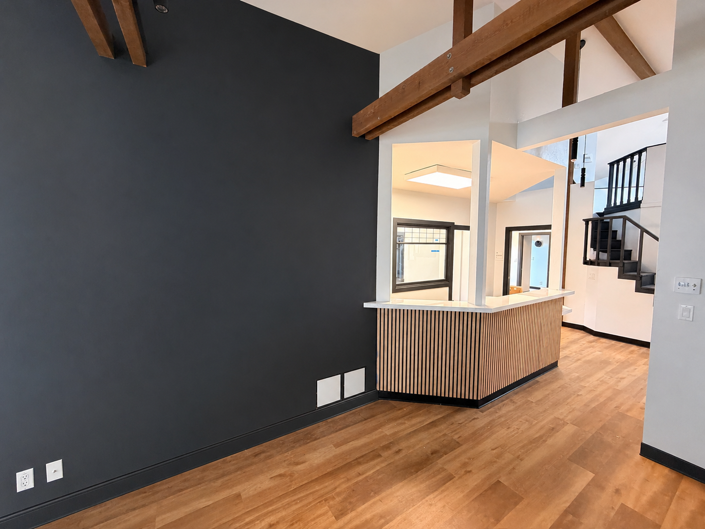 Interior view of a modern home with a dark accent wall, wooden flooring, and an open kitchen area featuring a white countertop, vertical wooden slats, and large windows. A staircase with black railing is visible in the background.