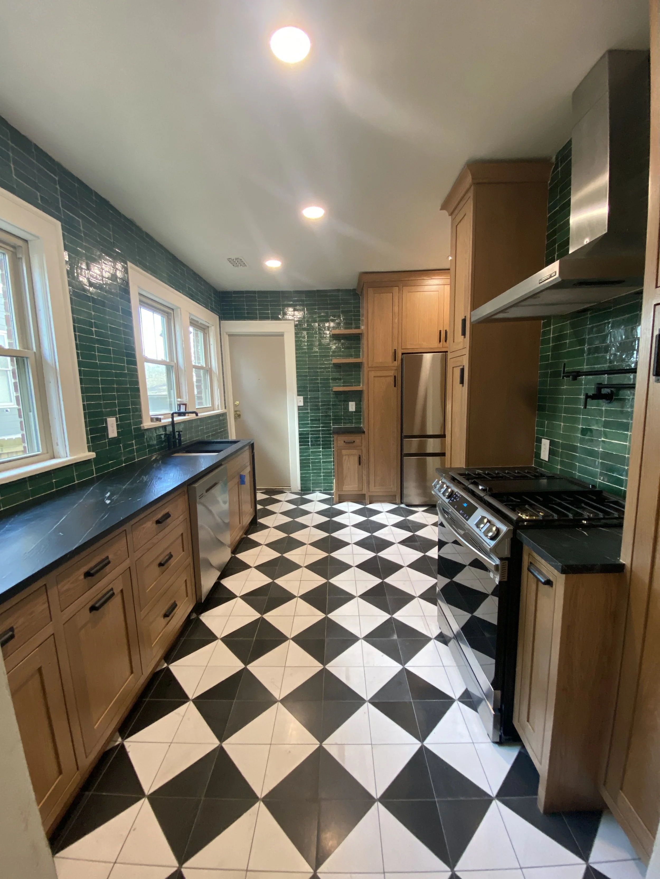 A kitchen with black and white checkered floor tiles, green tiled walls, wooden cabinets, and modern stainless steel appliances including a refrigerator and a stove.