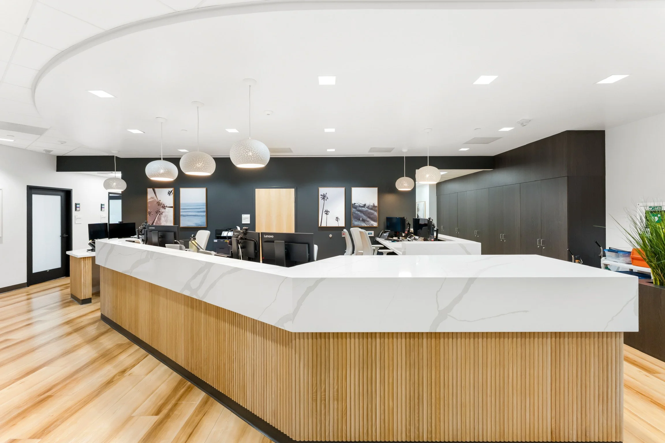 Modern office reception area with white marble counter, hanging pendant lights, black accent wall with framed pictures of a beach and palm trees, and wood flooring.