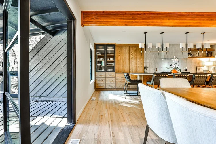 Interior of a modern dining area with a view to the kitchen, featuring wooden accents, a sliding glass door opening onto a deck, and a light-colored hardwood floor.