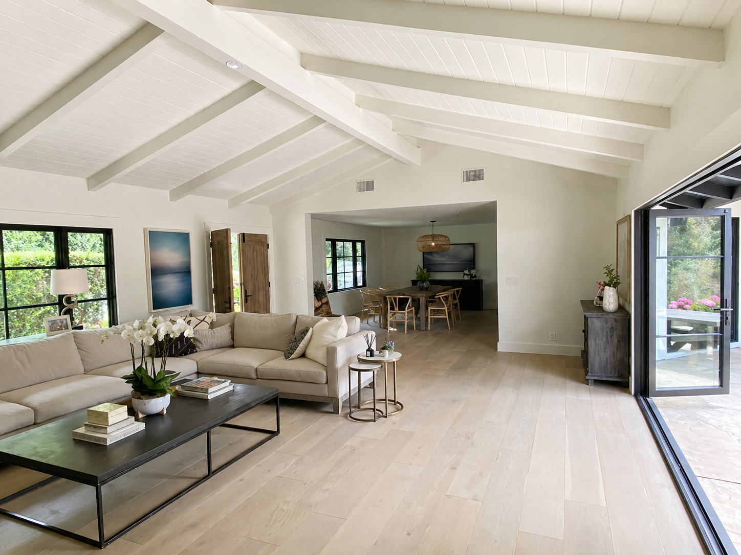 Open living room with beige sectional sofa, black coffee table, white orchid plant, and skylight ceiling, leading to a dining area with a wooden table and chairs, and sliding glass door to outdoor patio.