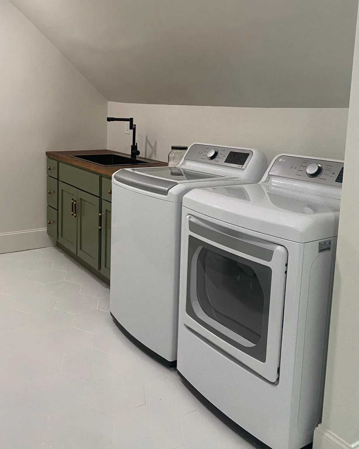 A laundry room with a front-loading washing machine and dryer, green cabinet with a wooden countertop, black faucet, and white hexagonal tile flooring.