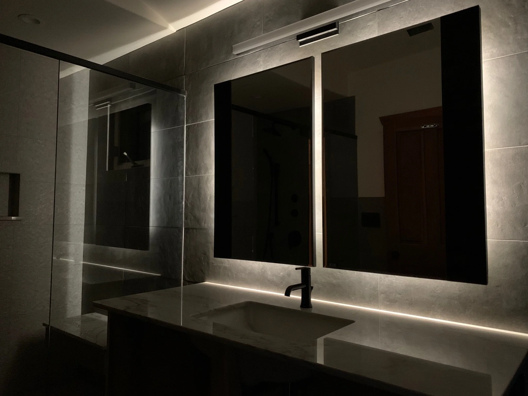 A dimly lit modern bathroom with gray tiled walls and floors, a rectangular mirror, and a dark wooden cabinet underneath the sink.