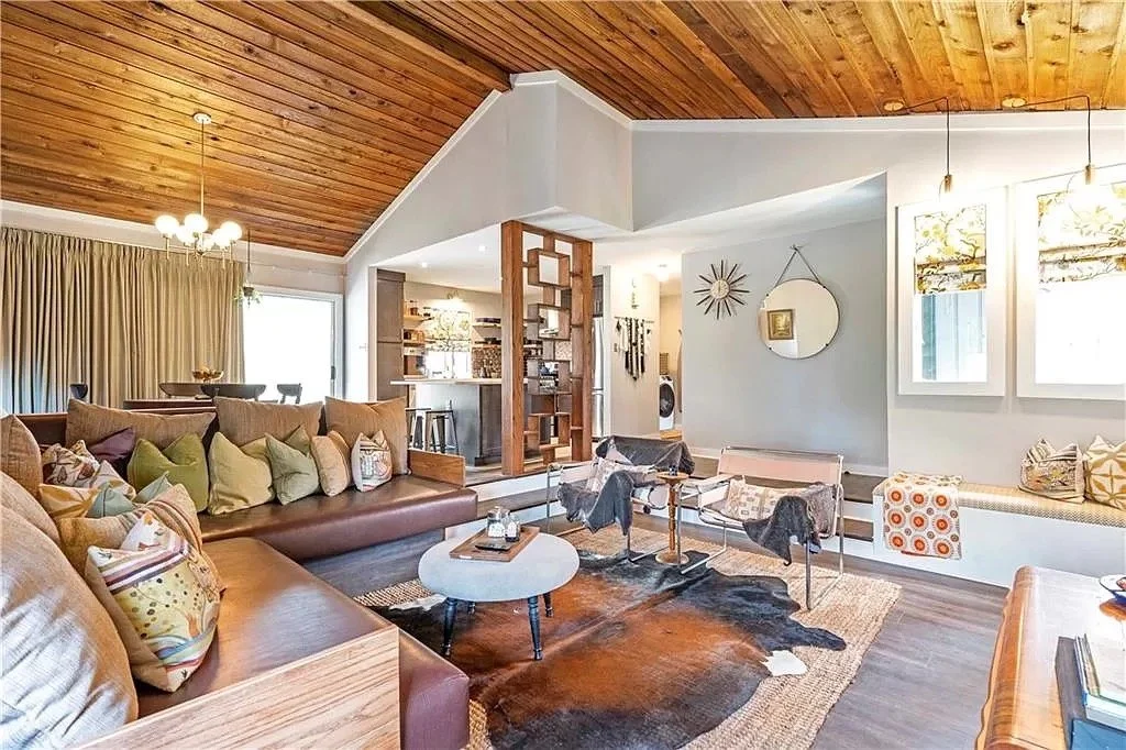 Living room with a wooden vaulted ceiling, large sectional sofa with colorful pillows, cowhide rug, small round coffee table, modern chairs, built-in window seat with cushions, decorative mirrors, and wall art.