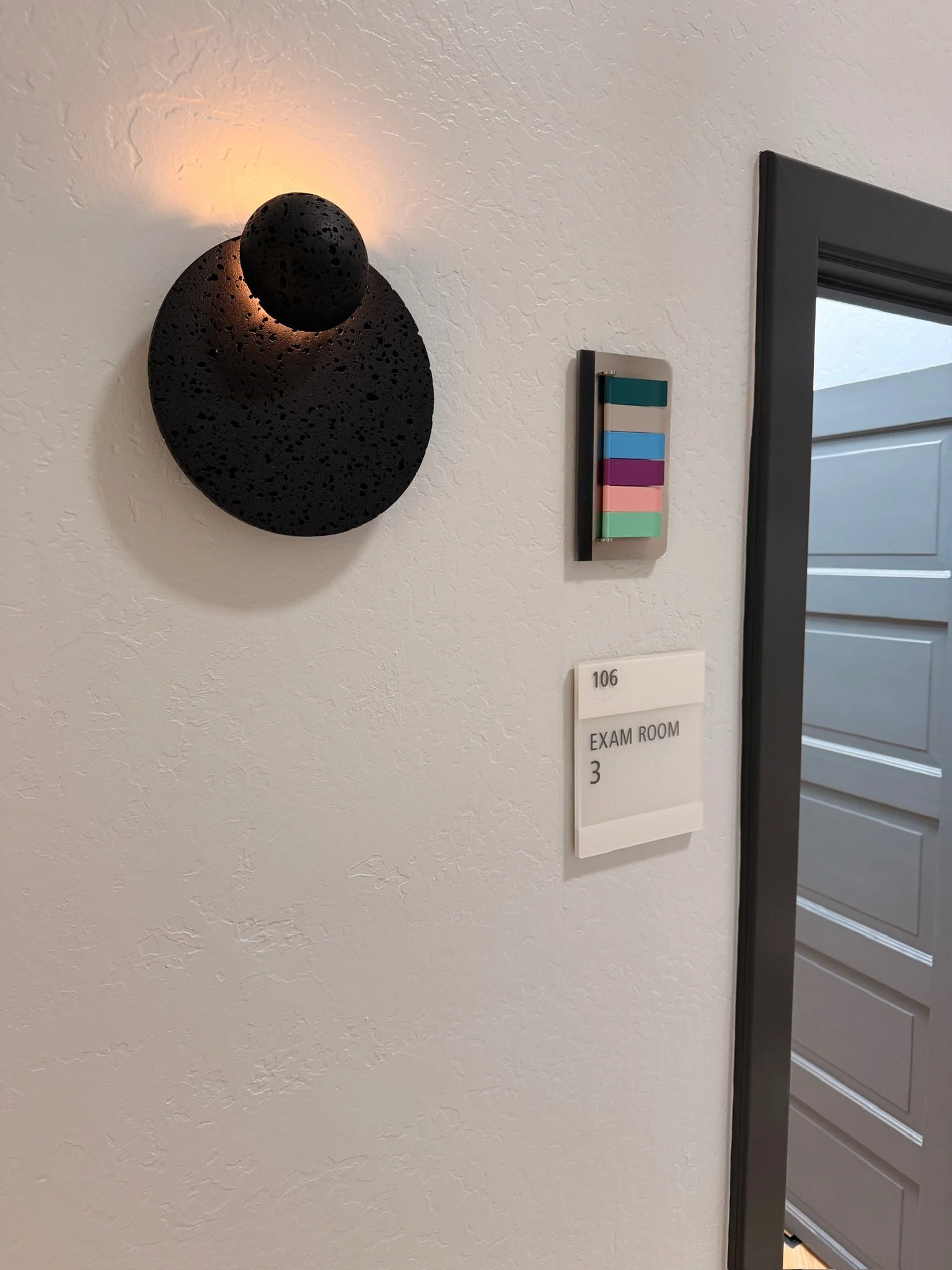 Decorative black wall sconce with a round light fixture on a white textured wall, next to a sign that reads "106 EXAM ROOM 3" and a door with horizontal paneling.