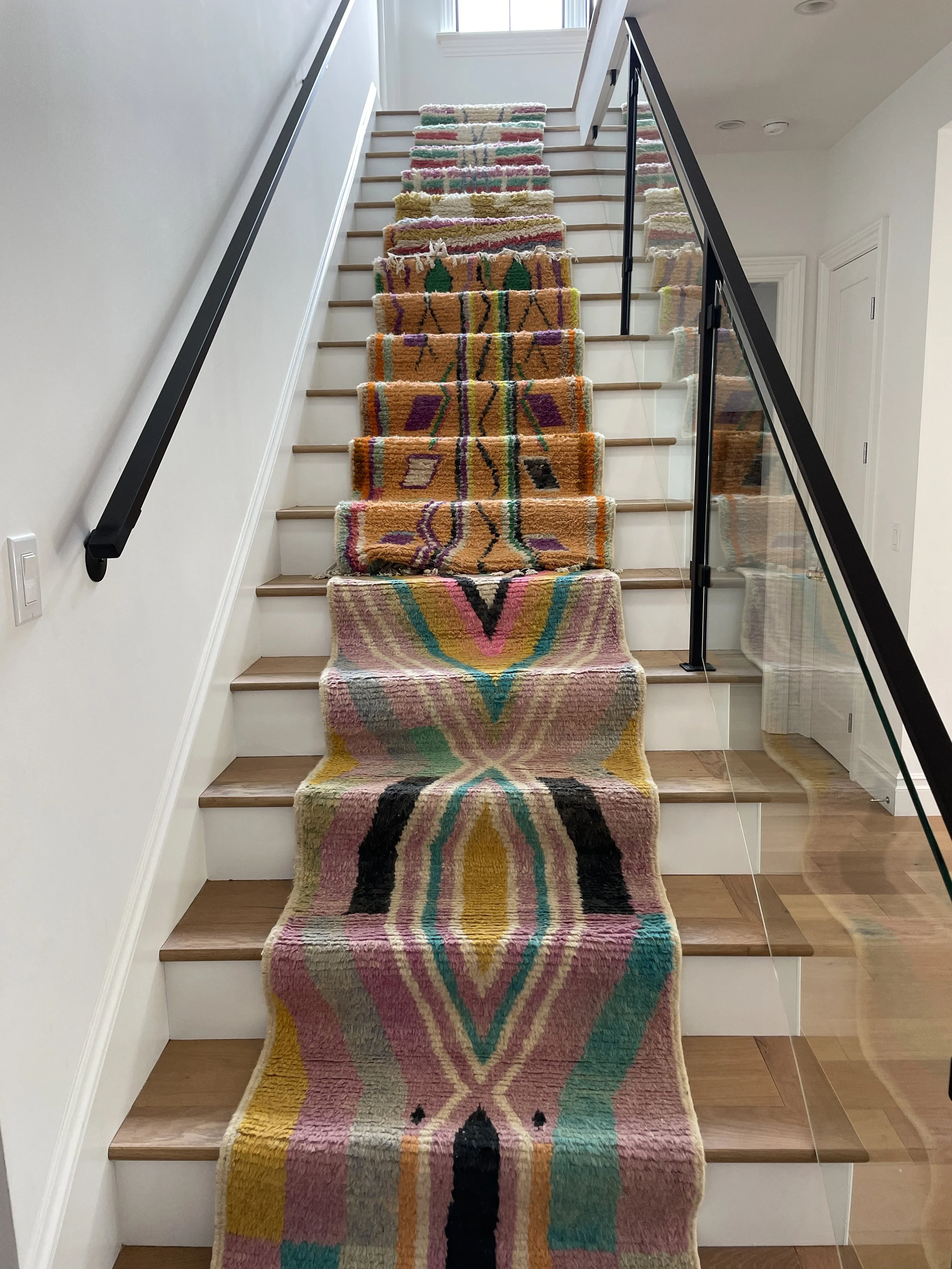Colorful abstract-patterned rug running down the center of a staircase with light wooden steps and a glass and metal handrail.