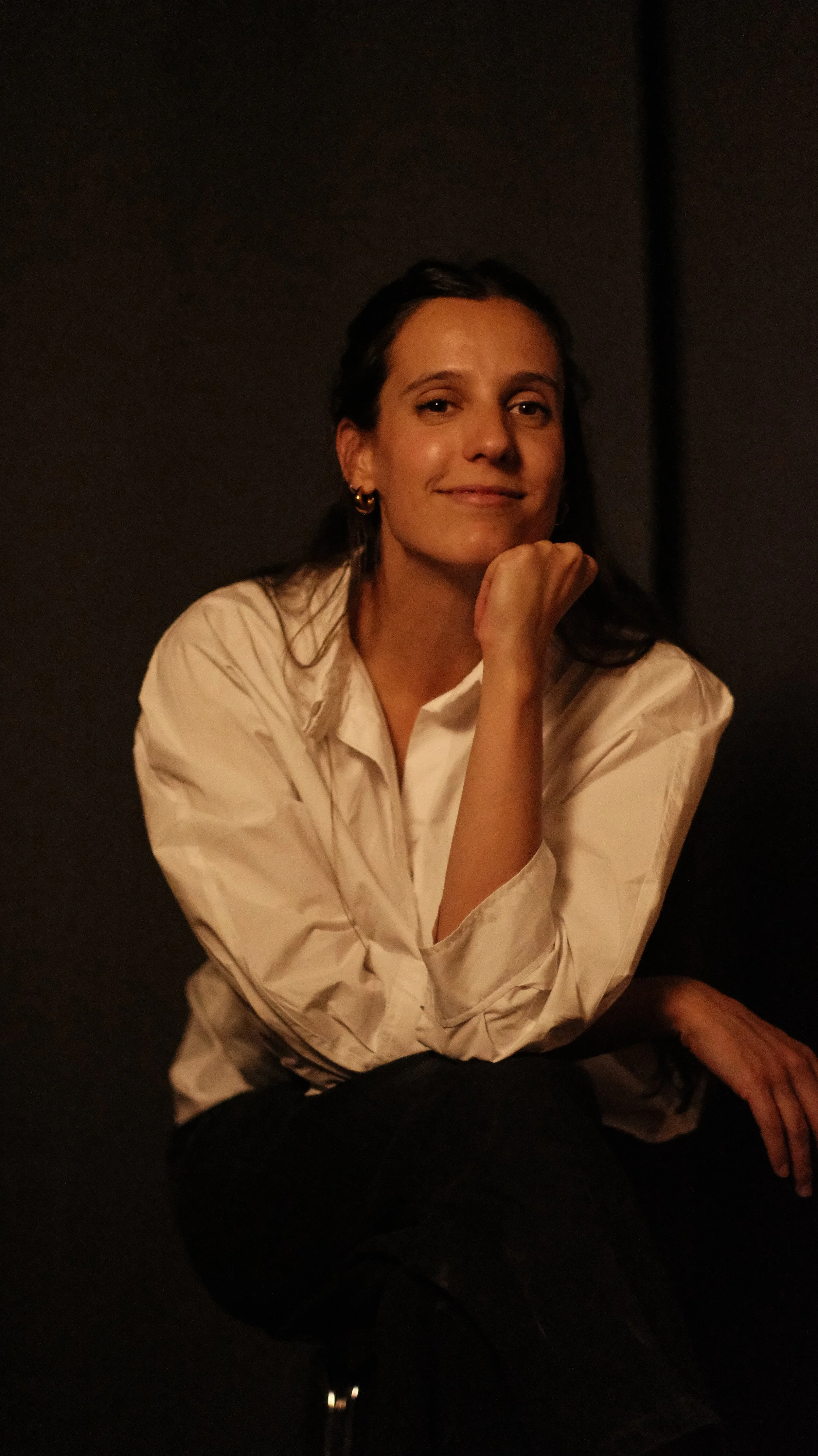 Mujer con blusa blanca, cabello oscuro, usando pendientes dorados, en ambiente oscuro, sonriendo y apoyando su rostro en su mano.