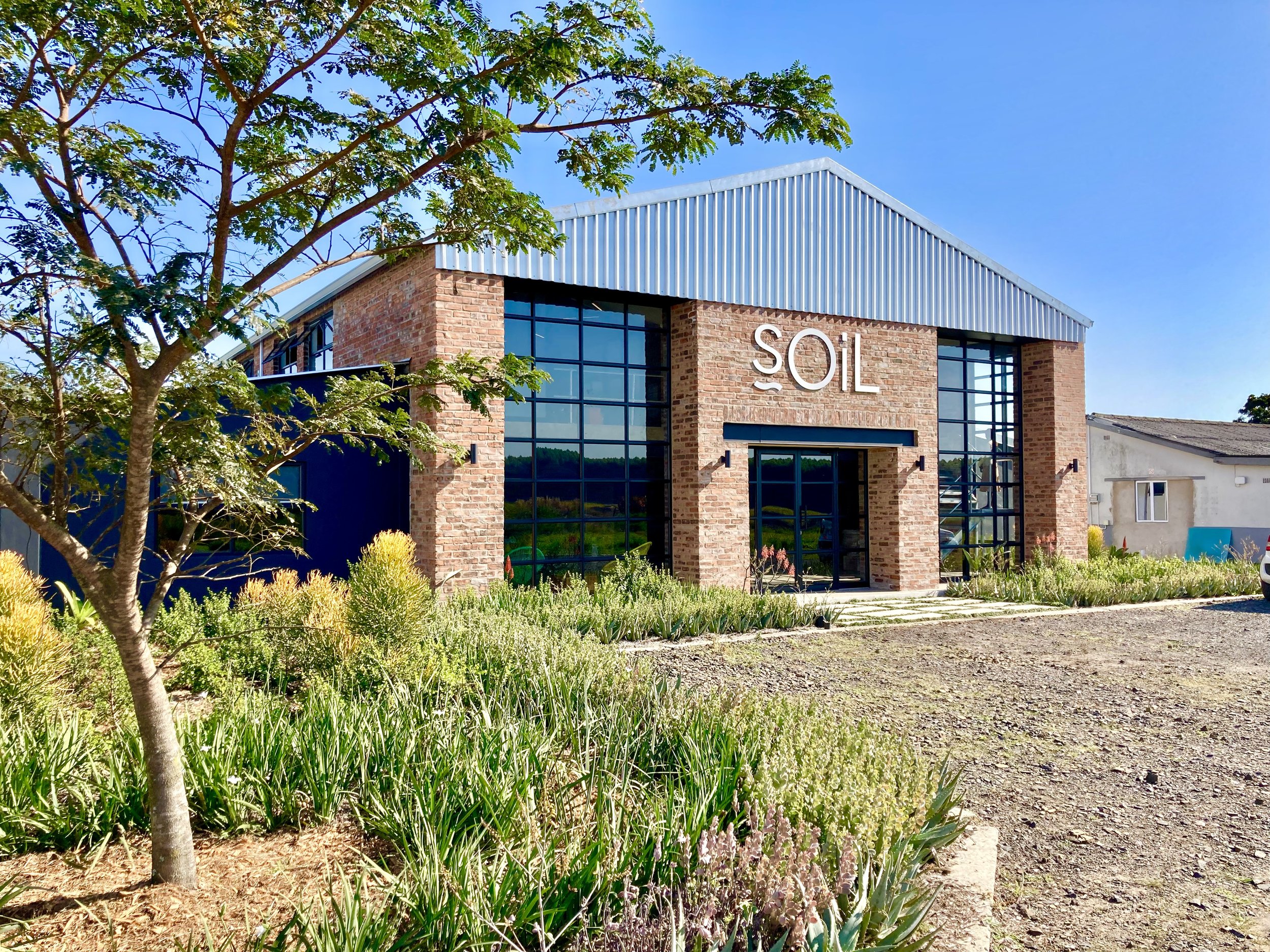 A modern brick building with large glass windows and the word 'SOIL' on the front, surrounded by green plants and trees under a clear blue sky.