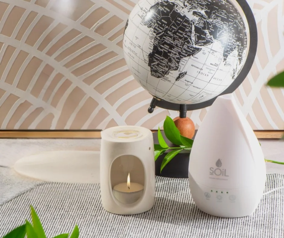 A globe, a white candle holder with a lit tea light, a white essential oil diffuser labeled 'SOiL', and some green leaves on a textured surface with a decorative background behind.