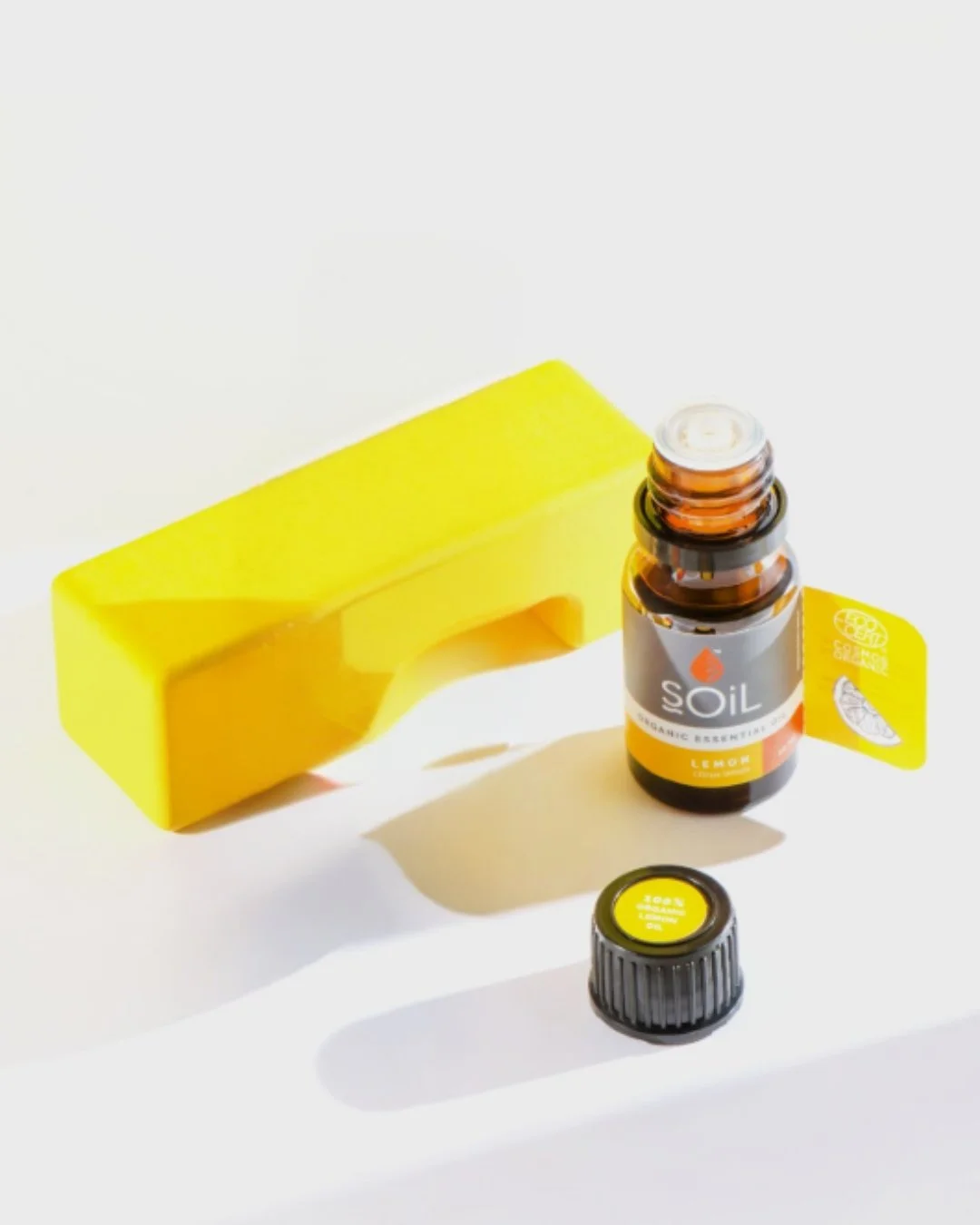 A small amber essential oil bottle labeled 'SOiL Organic Lemon Essential Oil' with a yellow cap, a yellow stand, and a yellow tag attached to the bottle, all on a white surface.
