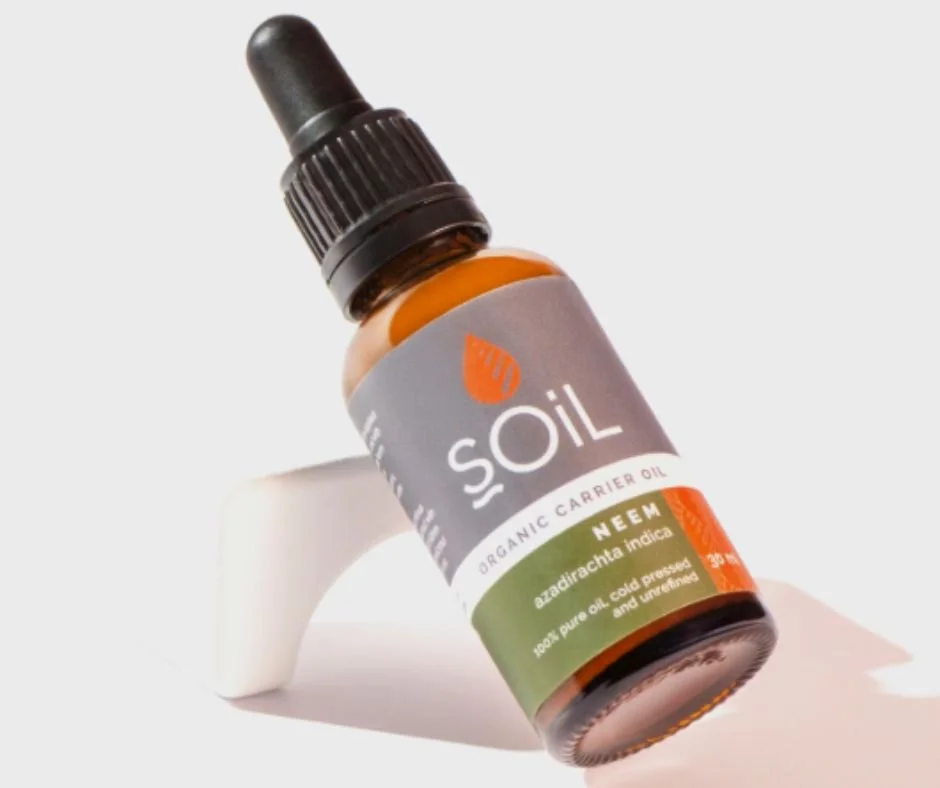 A small brown glass bottle of organic carrier oil labeled 'SOiL Organic Neem Carrier Oil', with a black dropper cap, lying on a white surface with product label details.