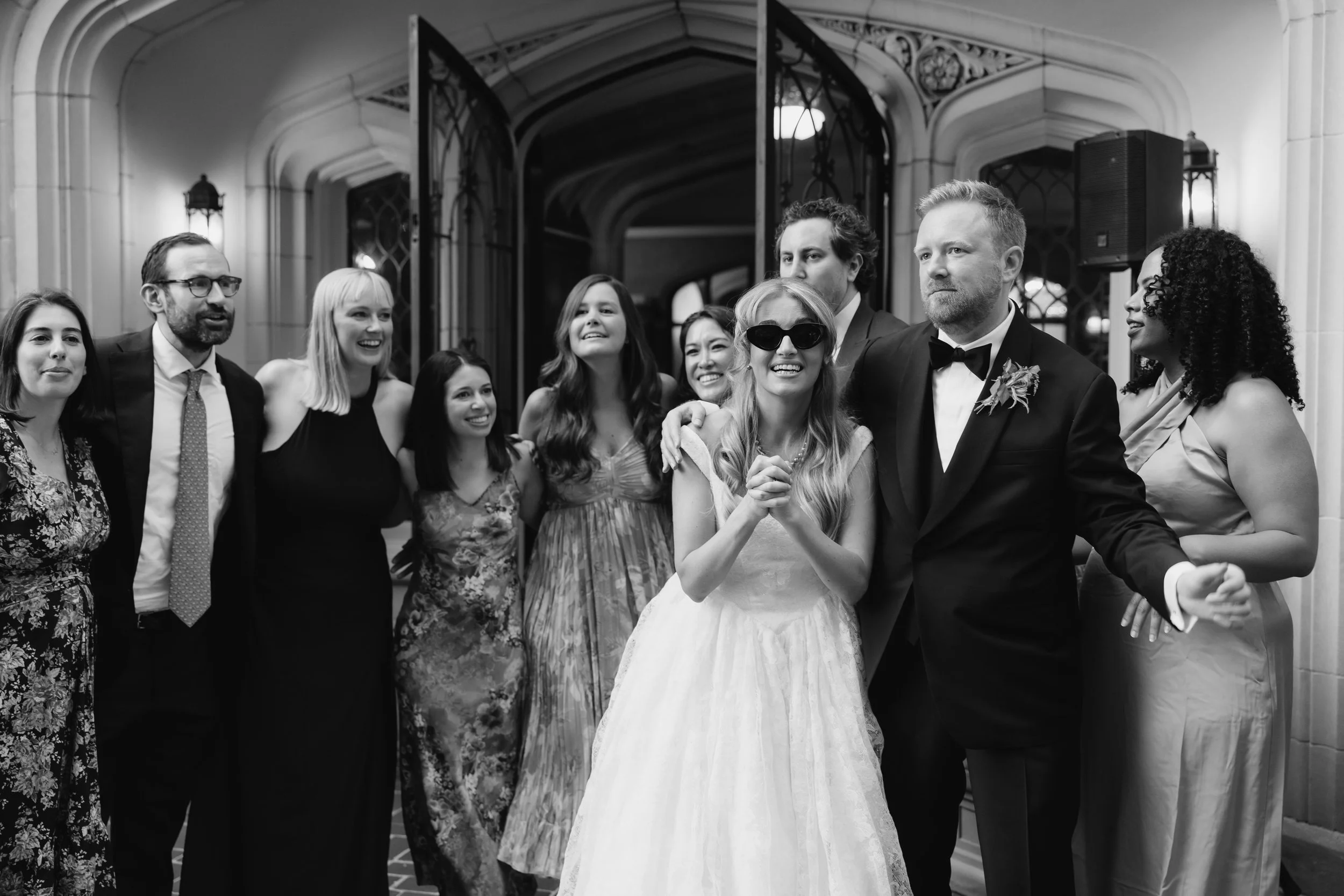 A group of people at a wedding celebration, with a bride in a white dress and sunglasses, and a groom in a tuxedo, posing happily with friends and family in an elegant indoor setting.