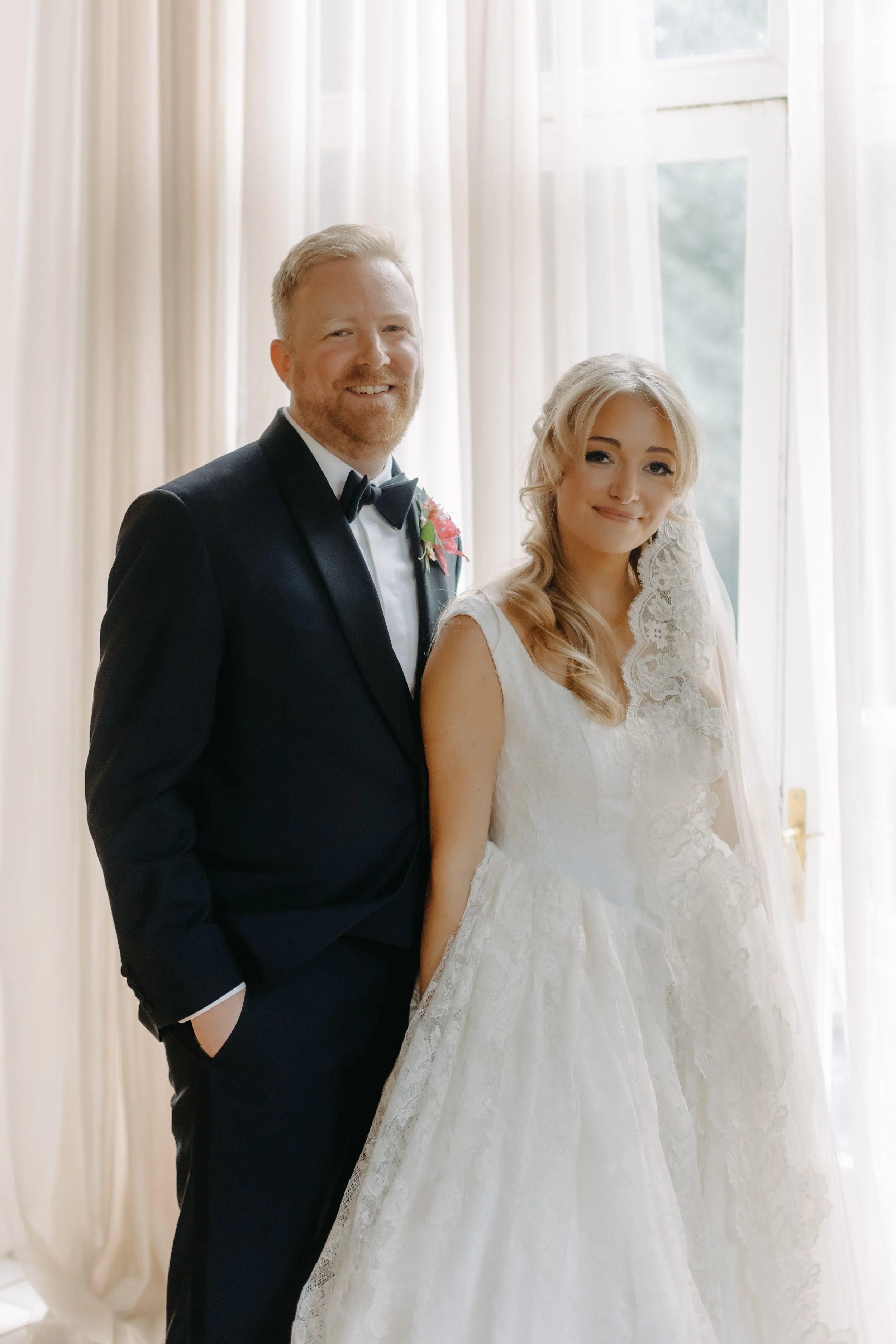 Bride and groom standing indoors in front of a bright window, smiling for a wedding photo. The groom wears a black tuxedo with a bow tie, and the bride wears a lace wedding gown with a veil.