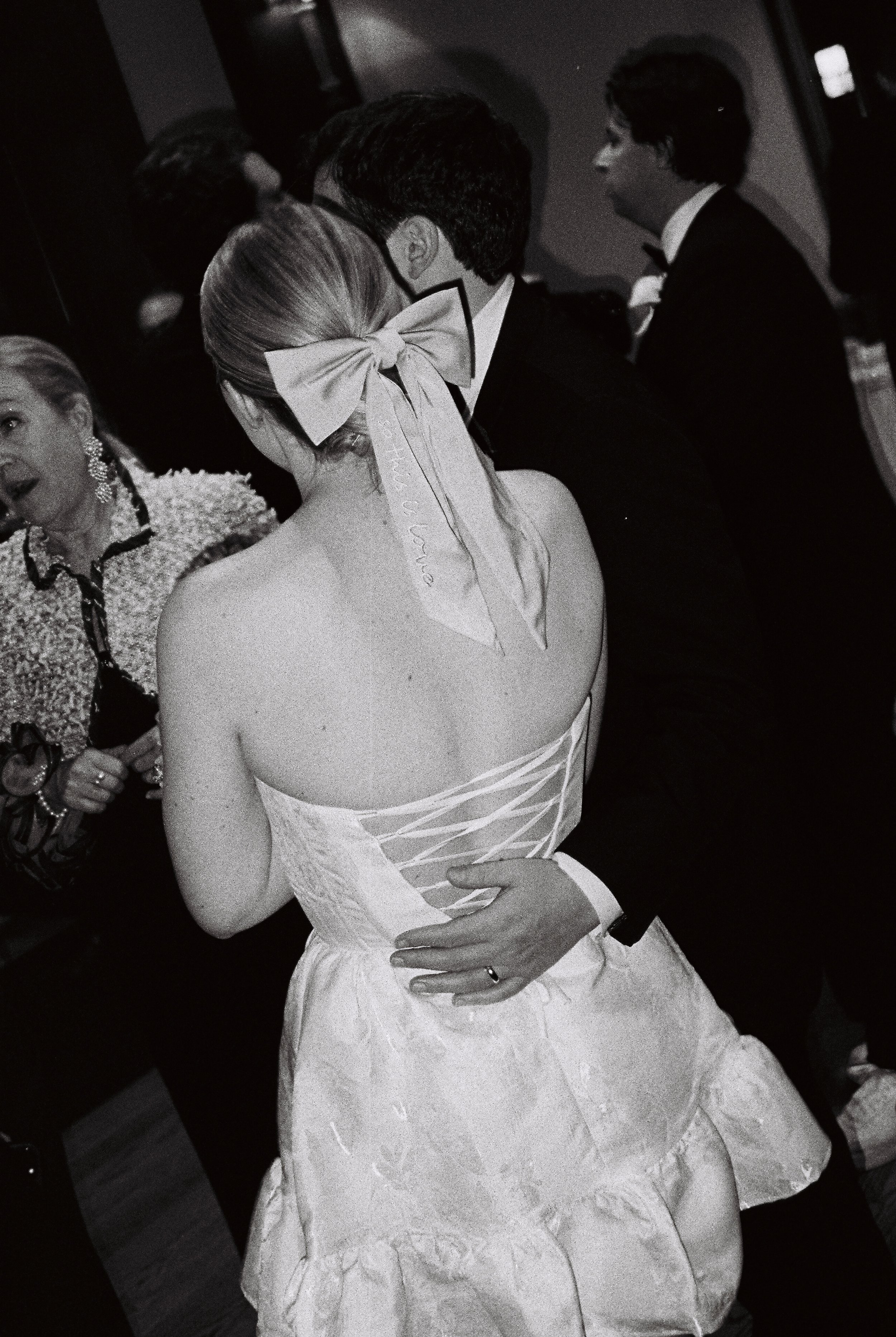 A woman in an off-shoulder dress with a large bow in her hair dancing with a man in a tuxedo at a formal event.