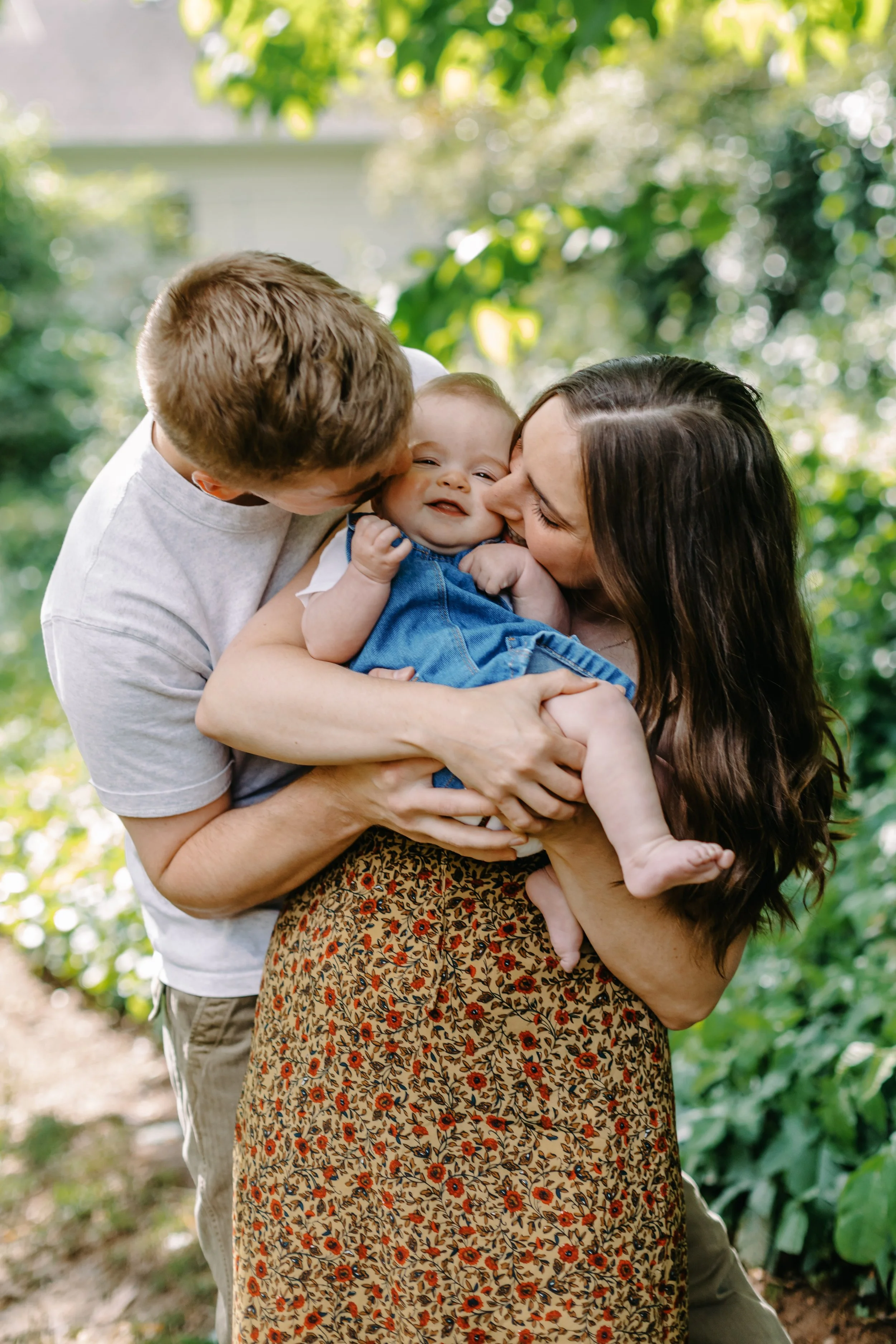 A family of three, a man, a woman, and a baby, outdoors in a lush green park, sharing a joyful kiss and holding the baby.