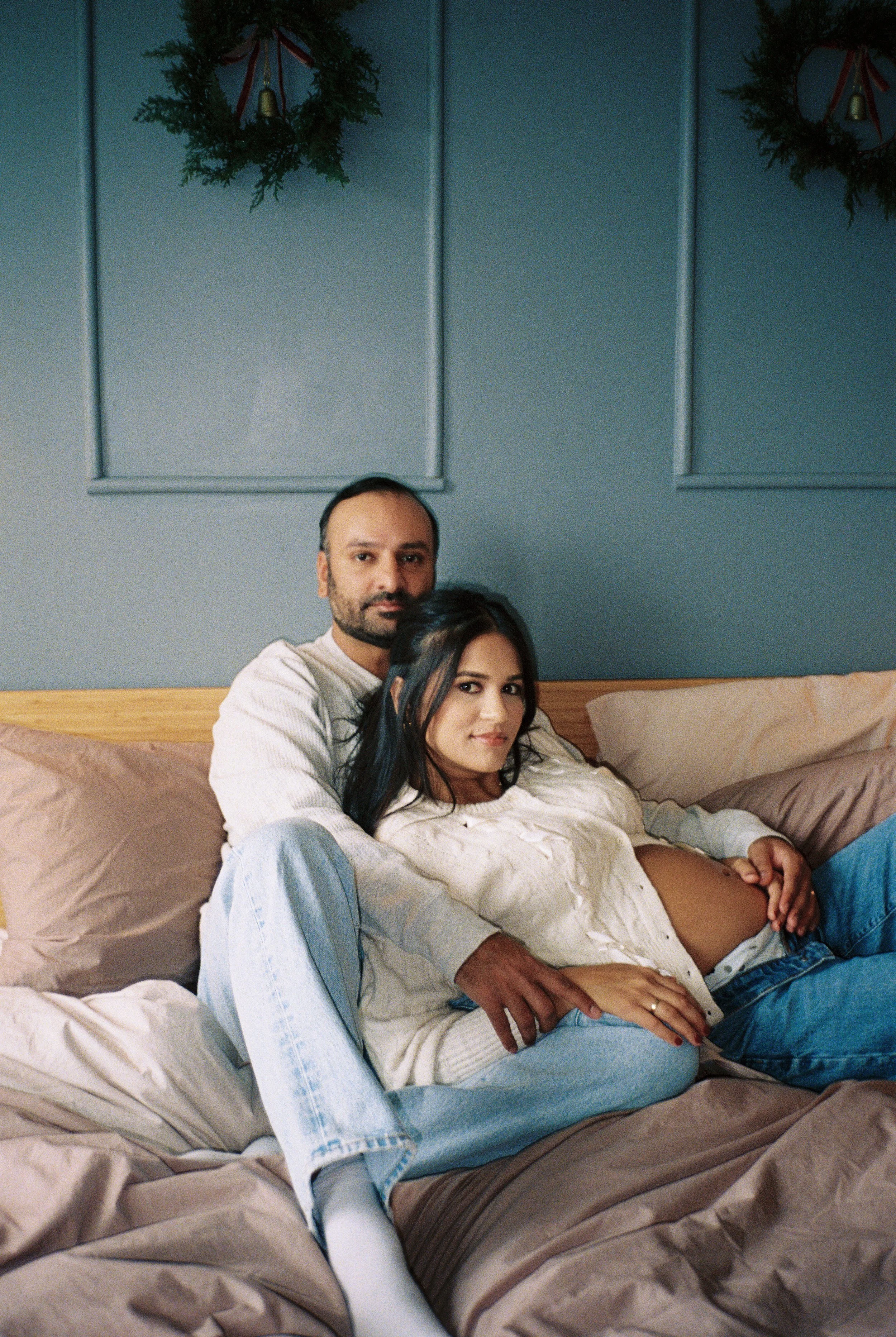 A couple sitting on a bed, with the woman showing a pregnant belly. The woman has dark hair, and the man has a beard. They are in a bedroom decorated with holiday-themed wreaths hanging on the wall.