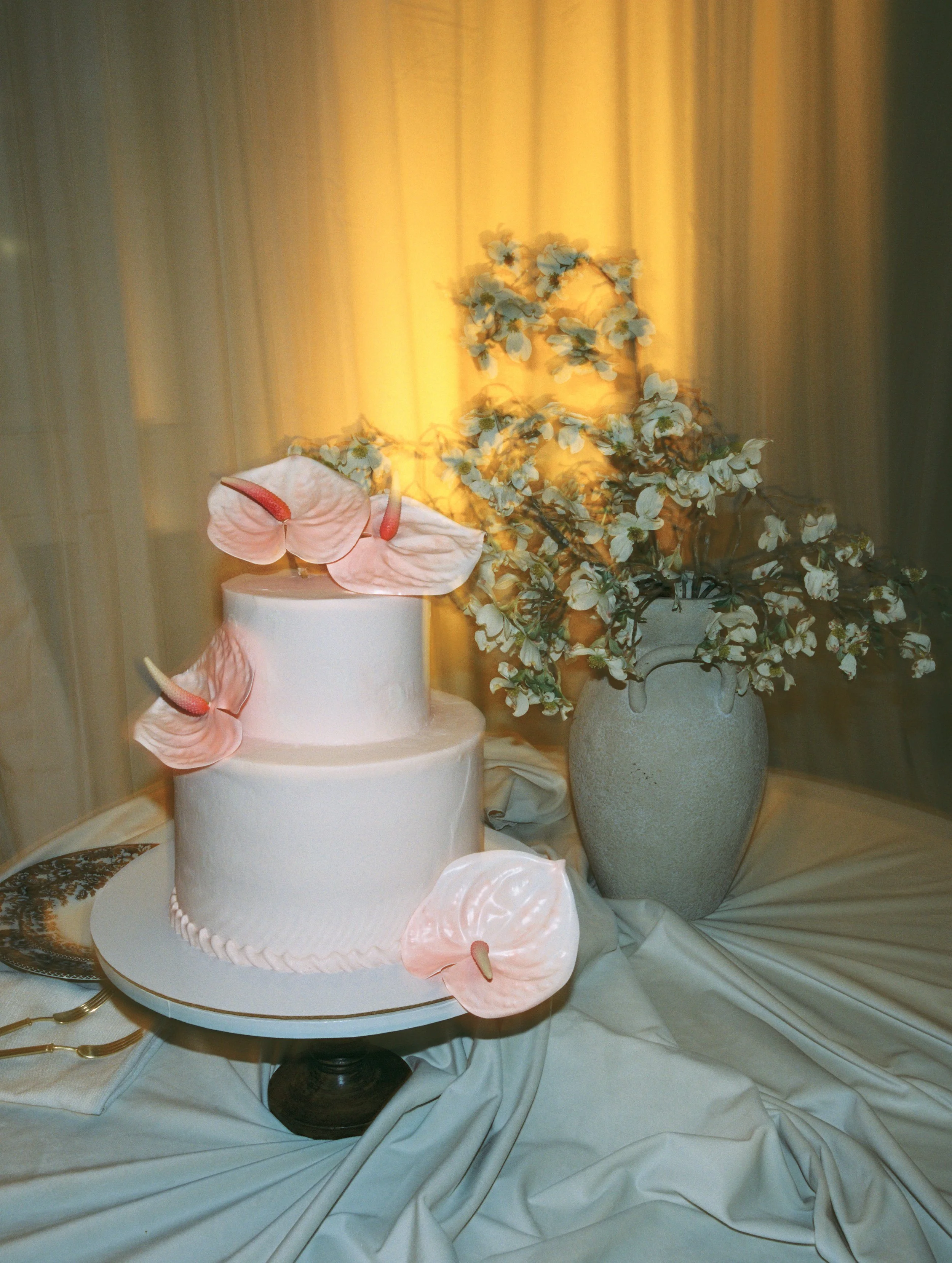 A two-tiered white wedding cake decorated with pink calla lilies, set on a black cake stand, beside a large vase of white flowers on a table with cream-colored drapery.