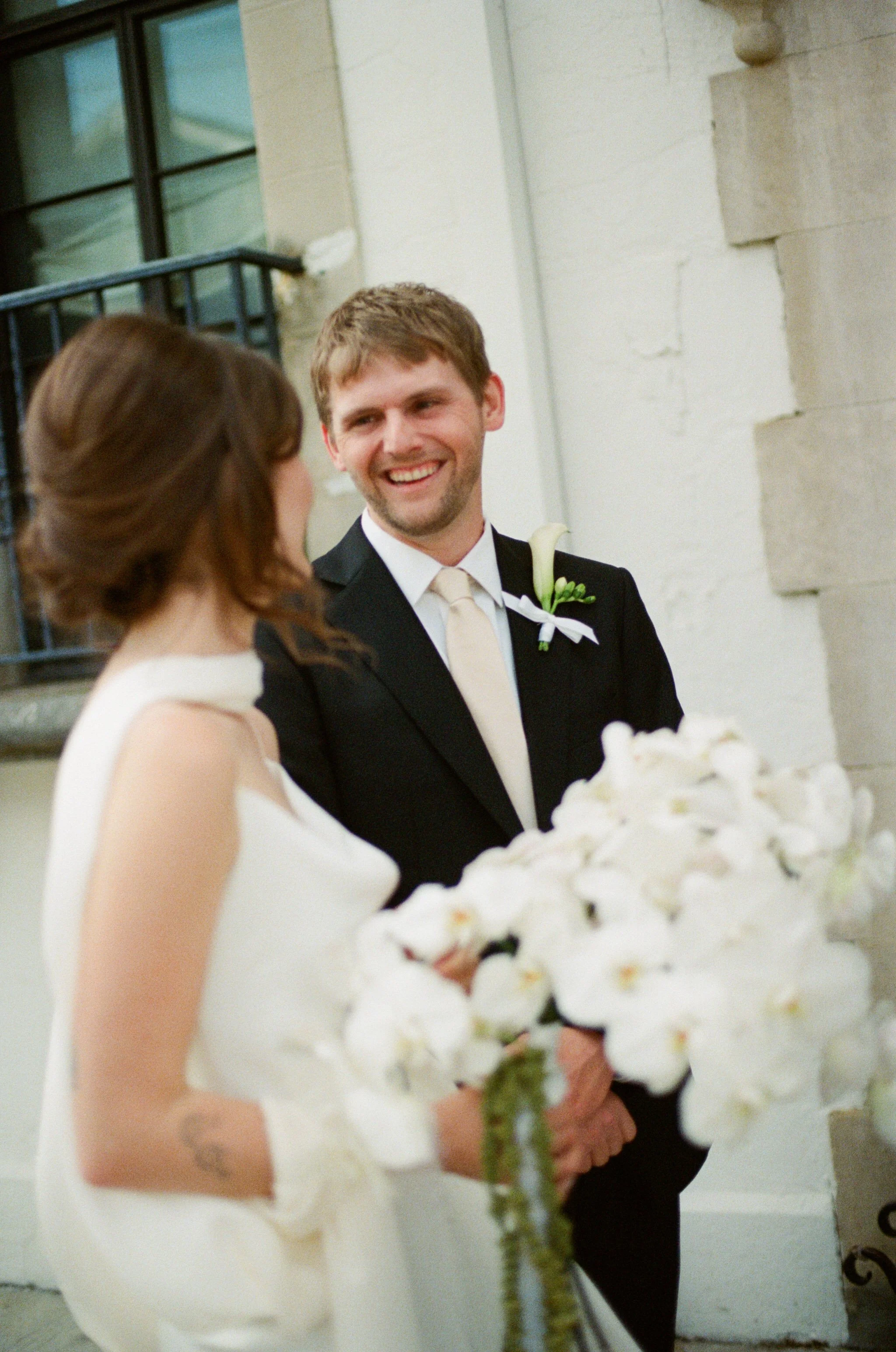 A bride and groom at their wedding, with the groom smiling and the bride holding a bouquet of white flowers, standing outdoors near a stone building.