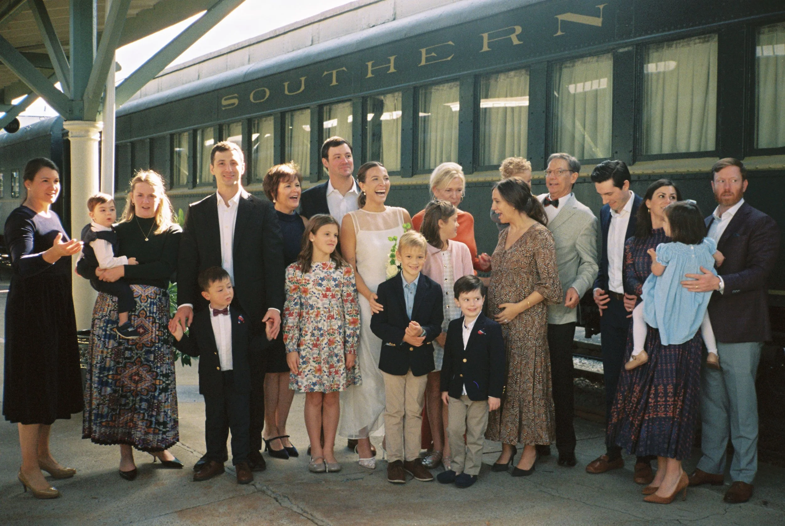 A group of people, including children and adults, standing on a train platform in front of a vintage train labeled 'Southern'. They appear to be dressed in formal or semi-formal attire, smiling and interacting.